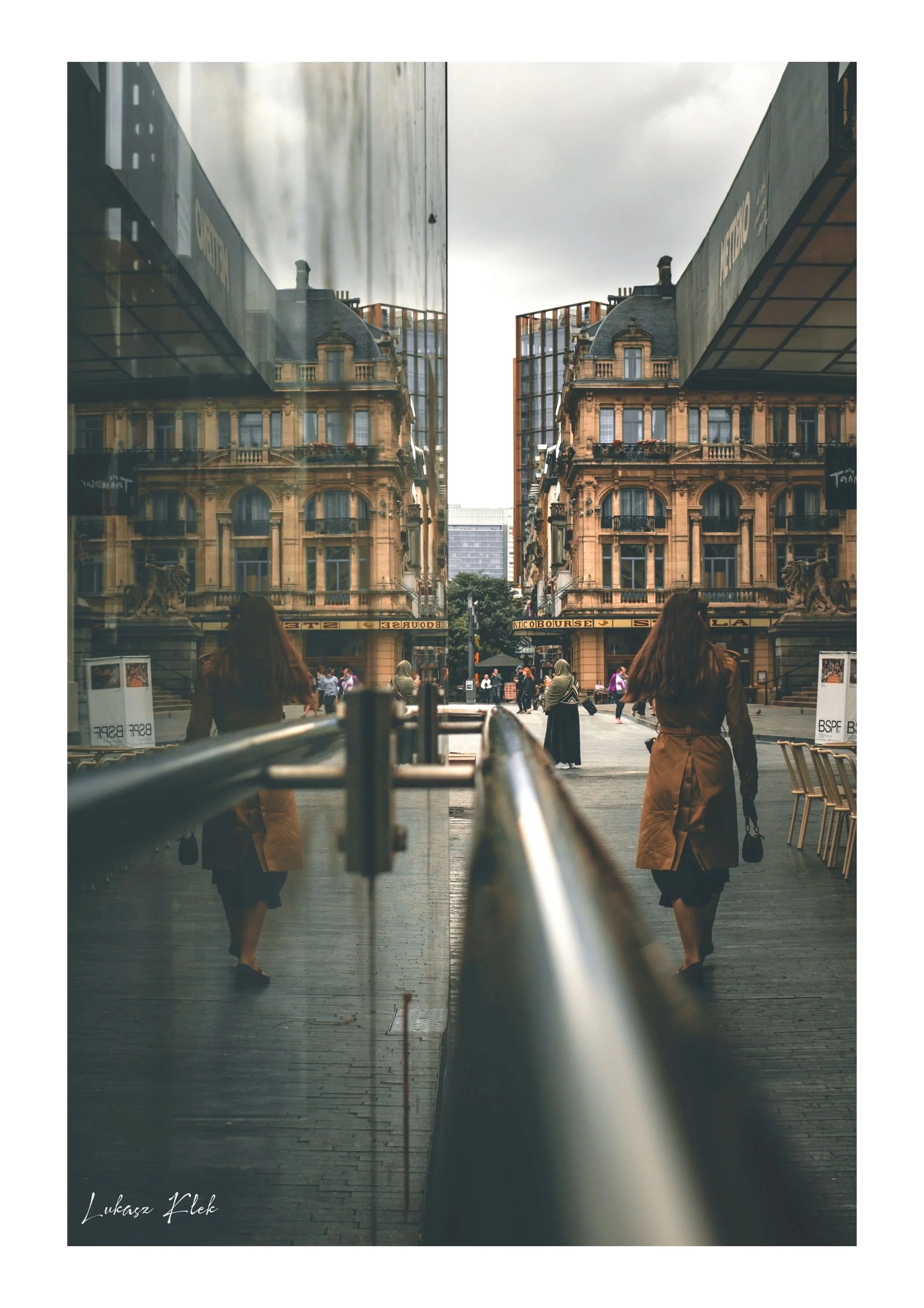 A city street scene reflected in a glass window, showing historic buildings with detailed architectural features, people walking, and outdoor seating.