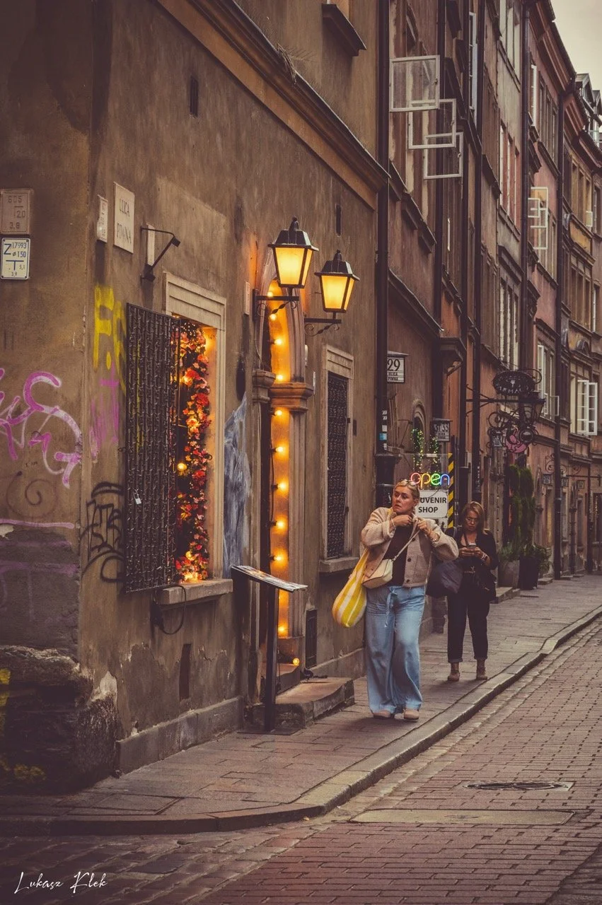 Two women walking on a cobblestone street in front of a building with warm yellow lights and graffiti, one woman is holding a yellow and white striped bag, and a neon sign that reads 'open' is visible.