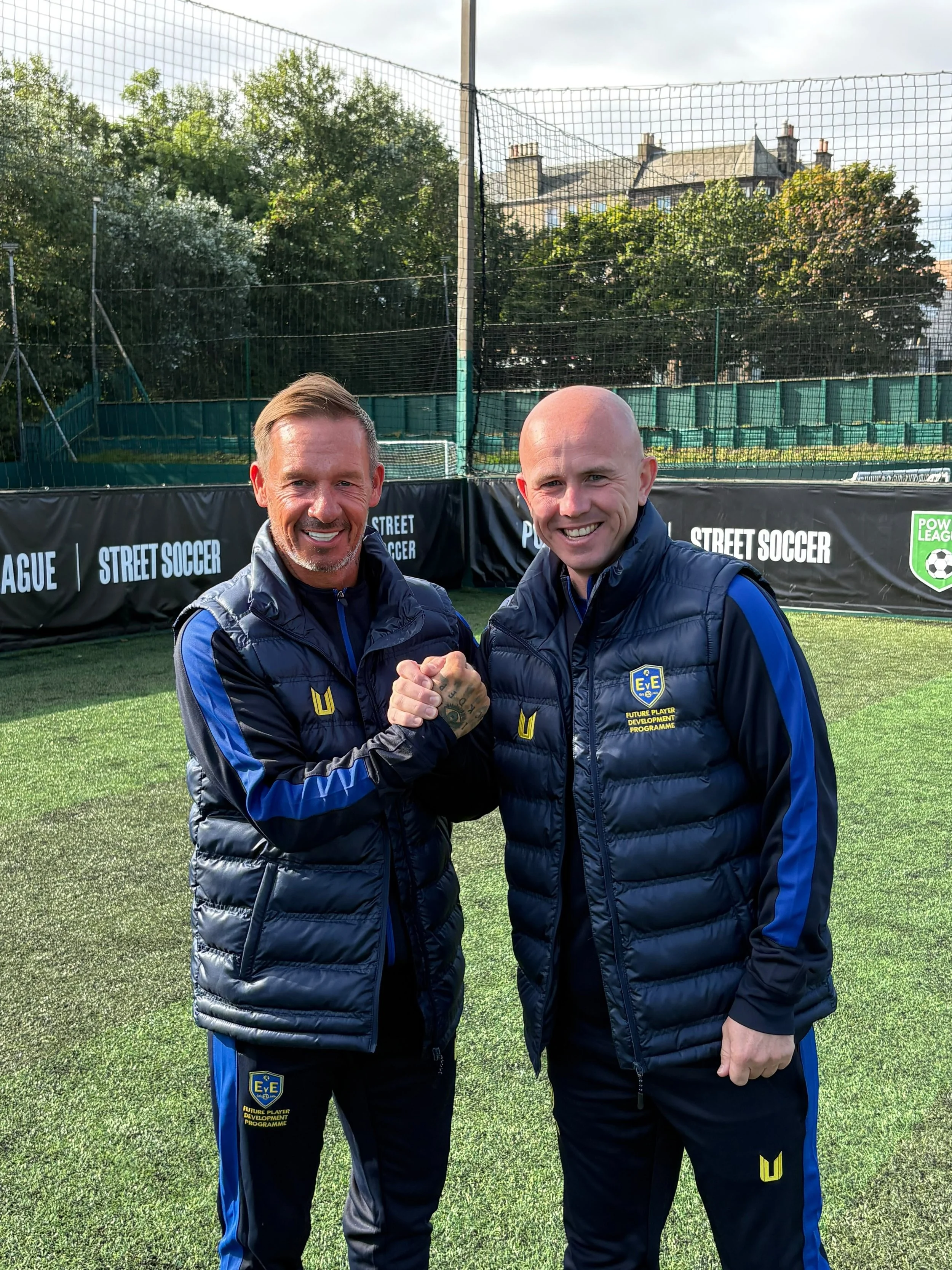 Two men in black and blue sports jackets shaking hands and smiling on a soccer field, with a goal net and green trees in the background.