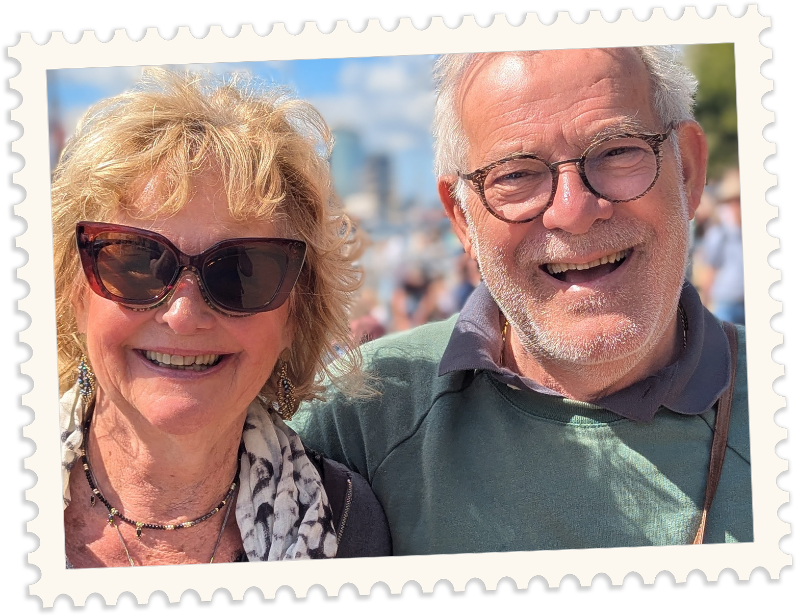 A smiling woman wearing large sunglasses, earrings, and a scarf, standing next to a smiling man with glasses and white hair, outdoors on a sunny day with a blurred crowd in the background.
