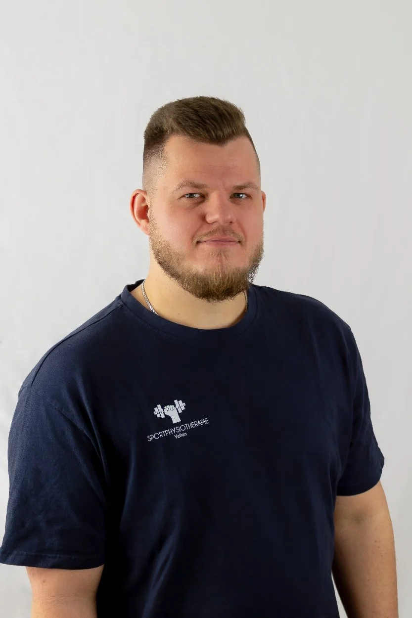 Porträt von Physiotherapeut Basti in der Praxis Sportphysiotherapie Velten, trägt ein Praxisshirt mit Logo vor einer grauen Wand.