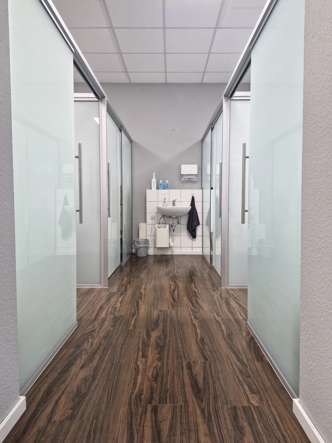 Heller, moderner Flur der Physiotherapie-Praxis in Velten mit Holzoptik-Laminat, abgehenden Behandlungsräumen und einem Waschbecken am Ende.