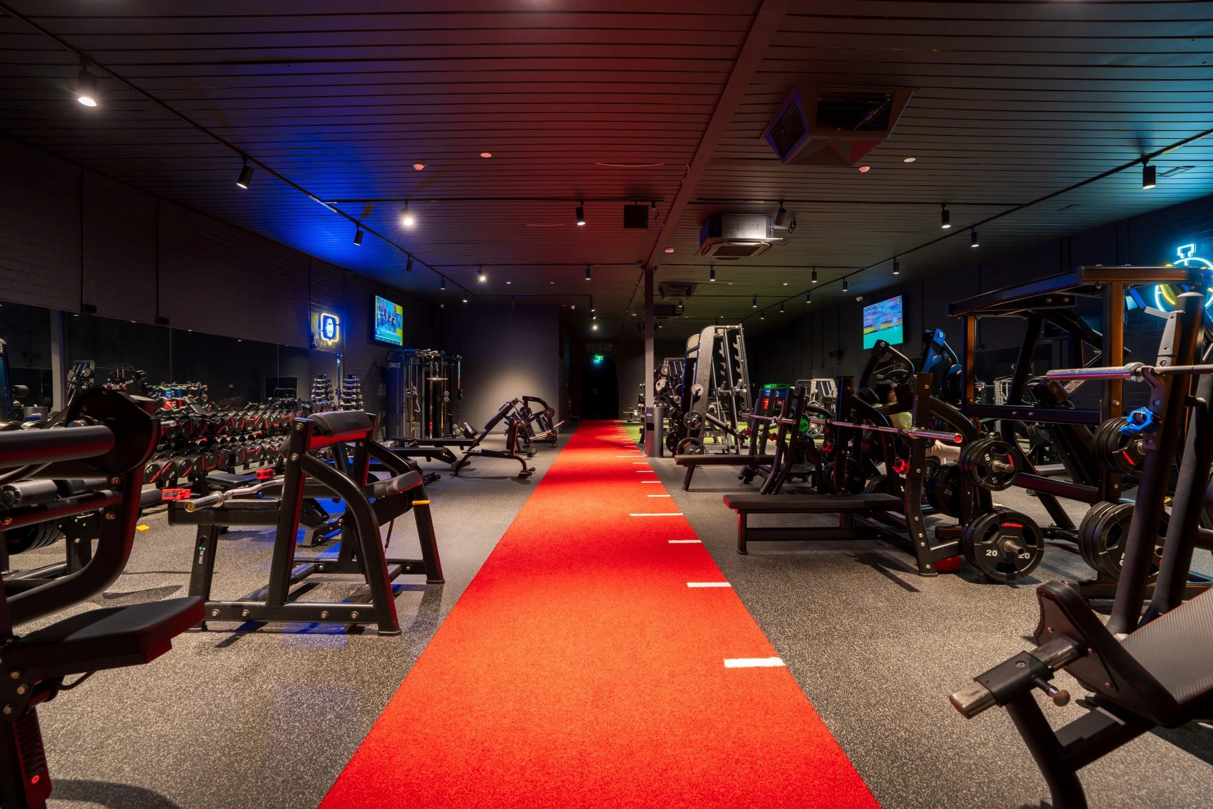Empty workout gym with weightlifting equipment, cardio machines, and a red carpeted lane in the center, illuminated by ceiling lights.