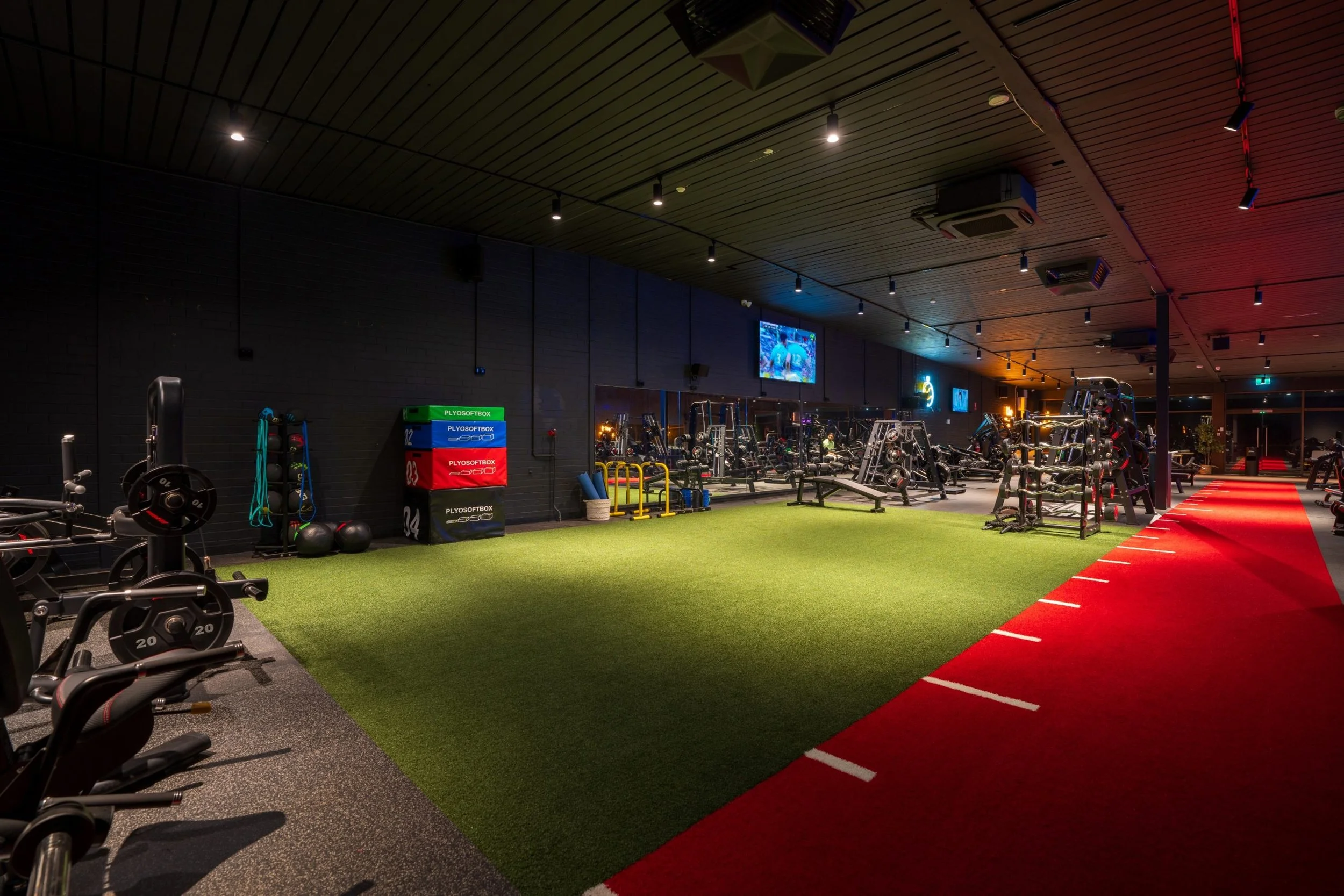 A brightly lit modern gym with various workout equipment, including weights, benches, and cardio machines. The gym has a red running track on the right side, black walls, a wooden ceiling, and multiple ceiling lights. A television screen is mounted o