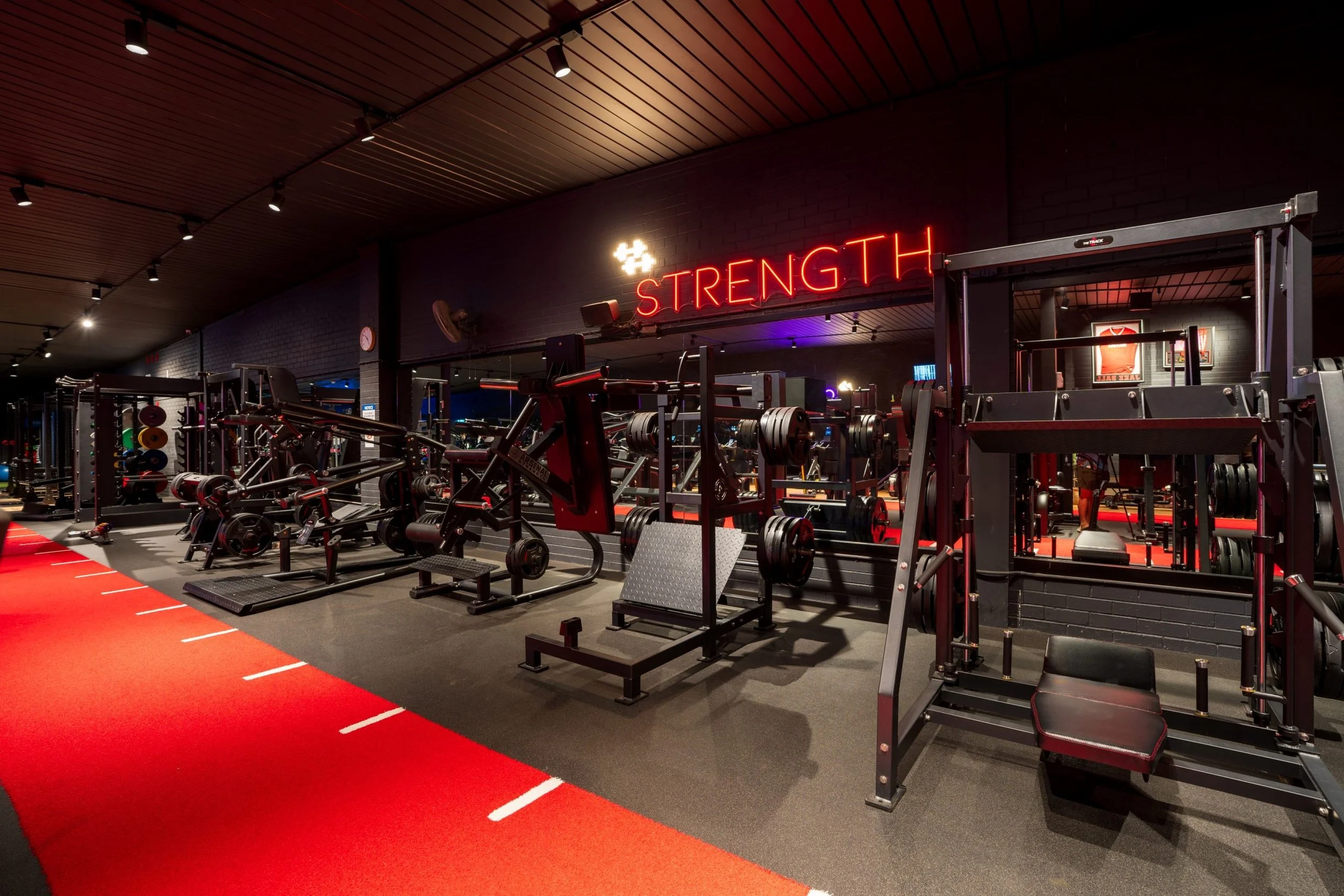 Gym with various workout equipment, weights, and machines, with a red sign that says 'Strength' on a black brick wall.