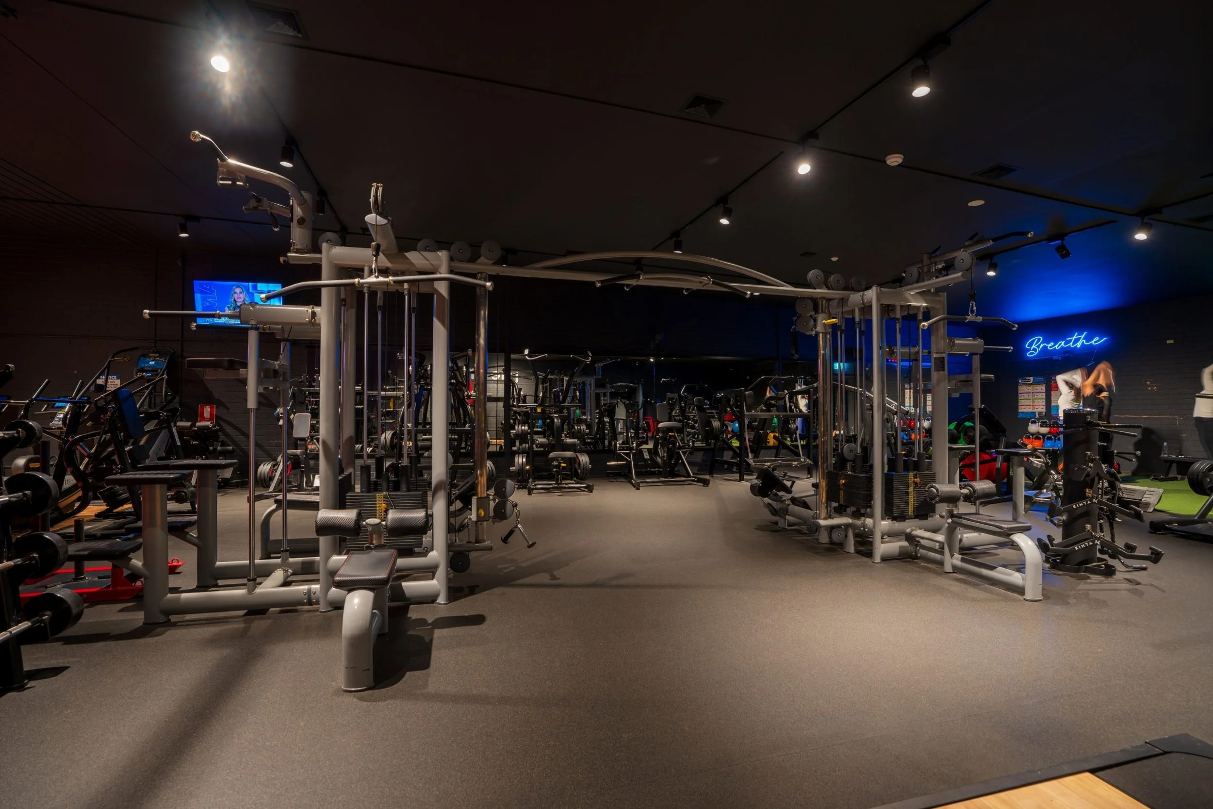 A spacious gym with various workout equipment including weight machines, free weights, and cardio machines, illuminated with ceiling spotlights and decorated with a blue neon sign that says 'Breathe'.