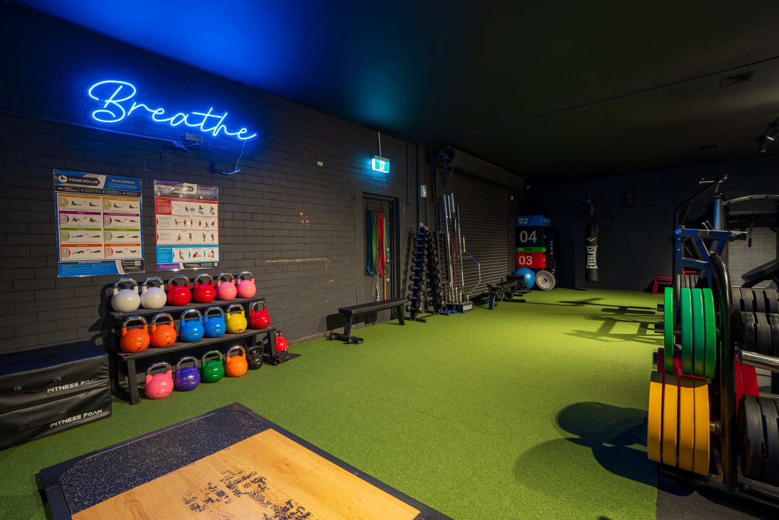 A gym with black brick walls illuminated by blue neon lighting that spells 'Breathe'. The gym has various fitness equipment, including colorful kettlebells, dumbbells, a punching bag, and exercise mats, with bright green artificial turf flooring.