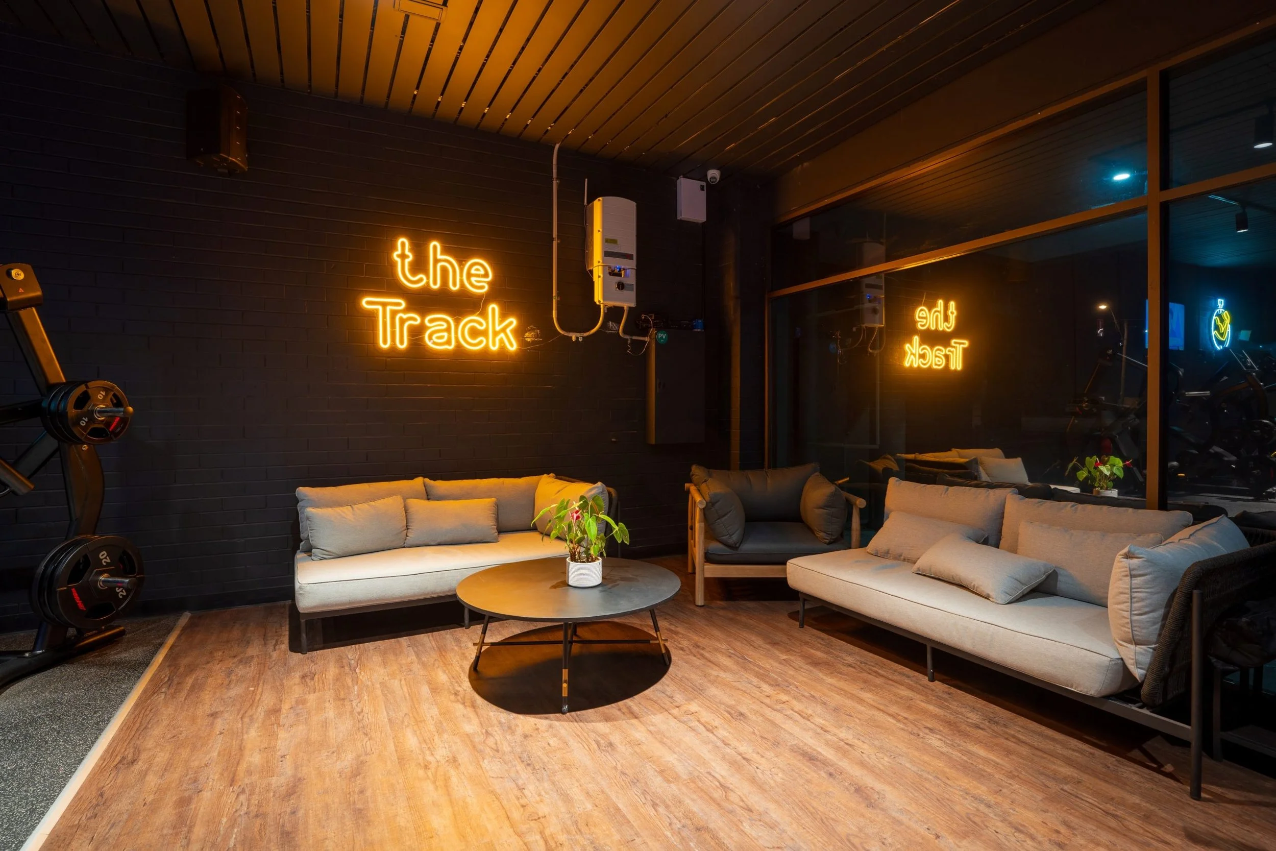 A cozy lounge area with beige sofas, a round coffee table with a potted plant, wood flooring, and a black brick wall. Neon signs read 'the Track' and 'the Track' reflected in the large glass window. Gym equipment and mirrors are visible in the backgr