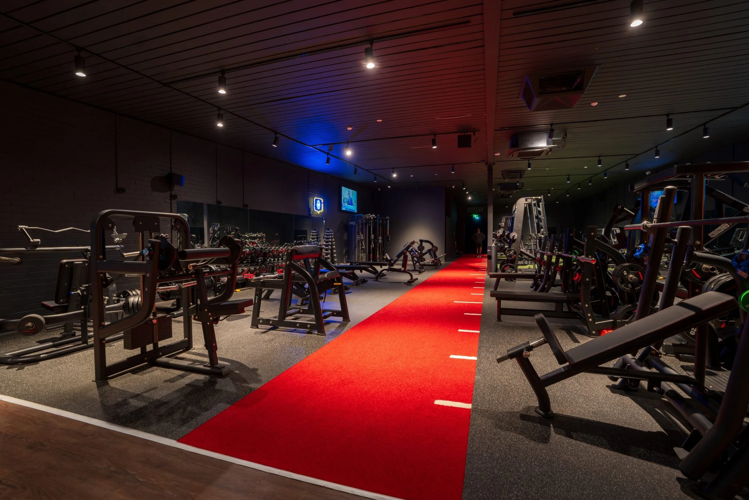 Interior view of a gym with various workout equipment, a red exercise mat in the center, and a red carpet running through the middle.