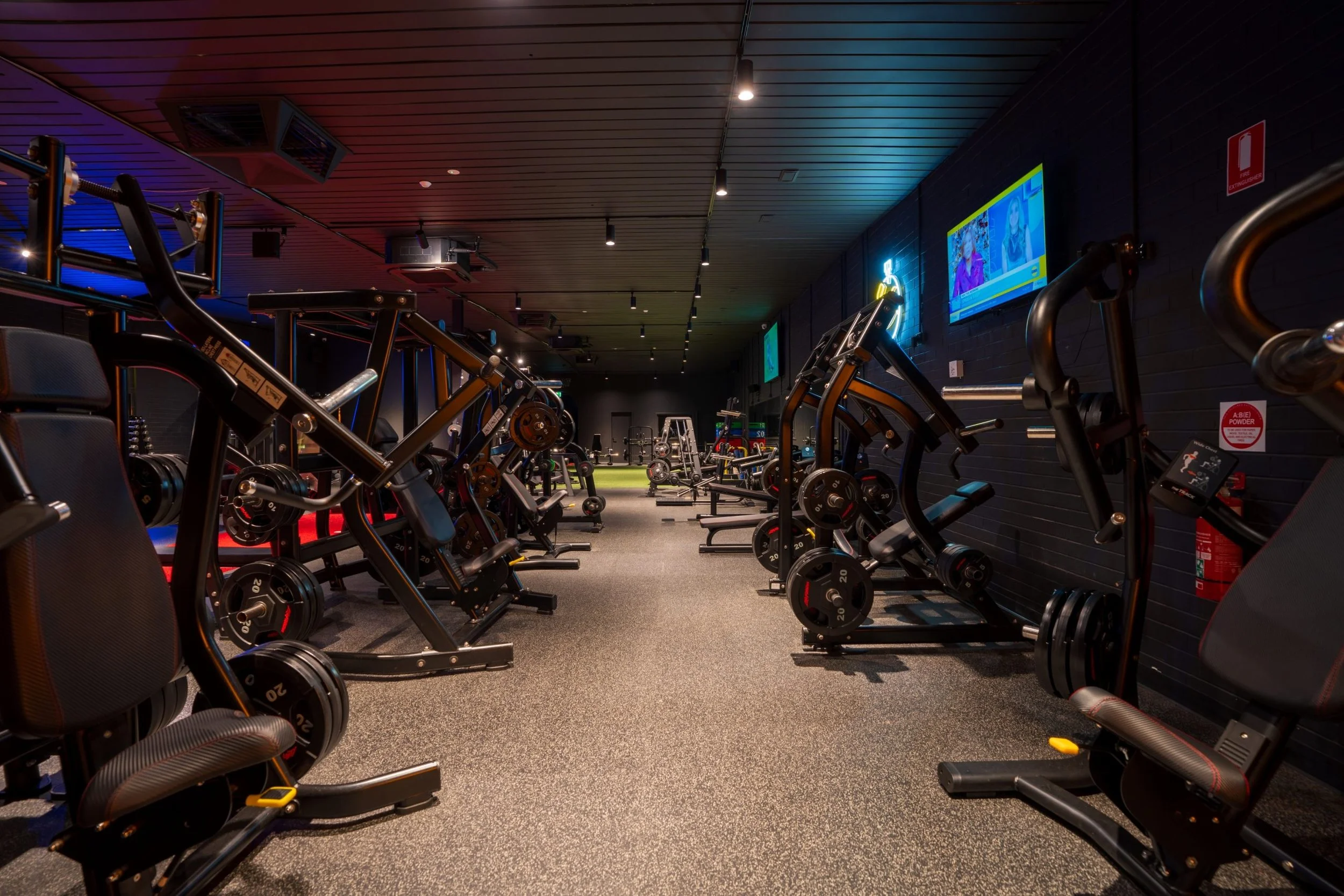 View of a modern gym with weightlifting equipment, exercise machines, and digital screens on the black wall, illuminated by ceiling lights.