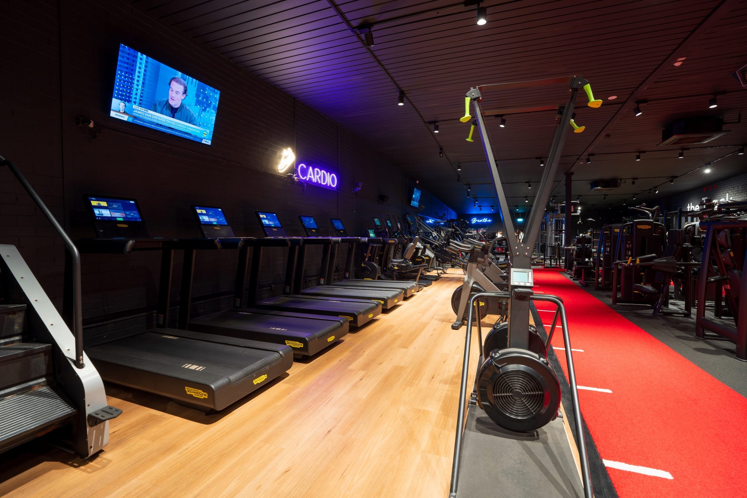 A gym with treadmills on a wooden floor, a red carpet, and various exercise equipment. A neon sign reads 'CARDIO', and multiple screens display workout data. The ceiling has black panels with small spotlights, and a TV shows a person speaking.