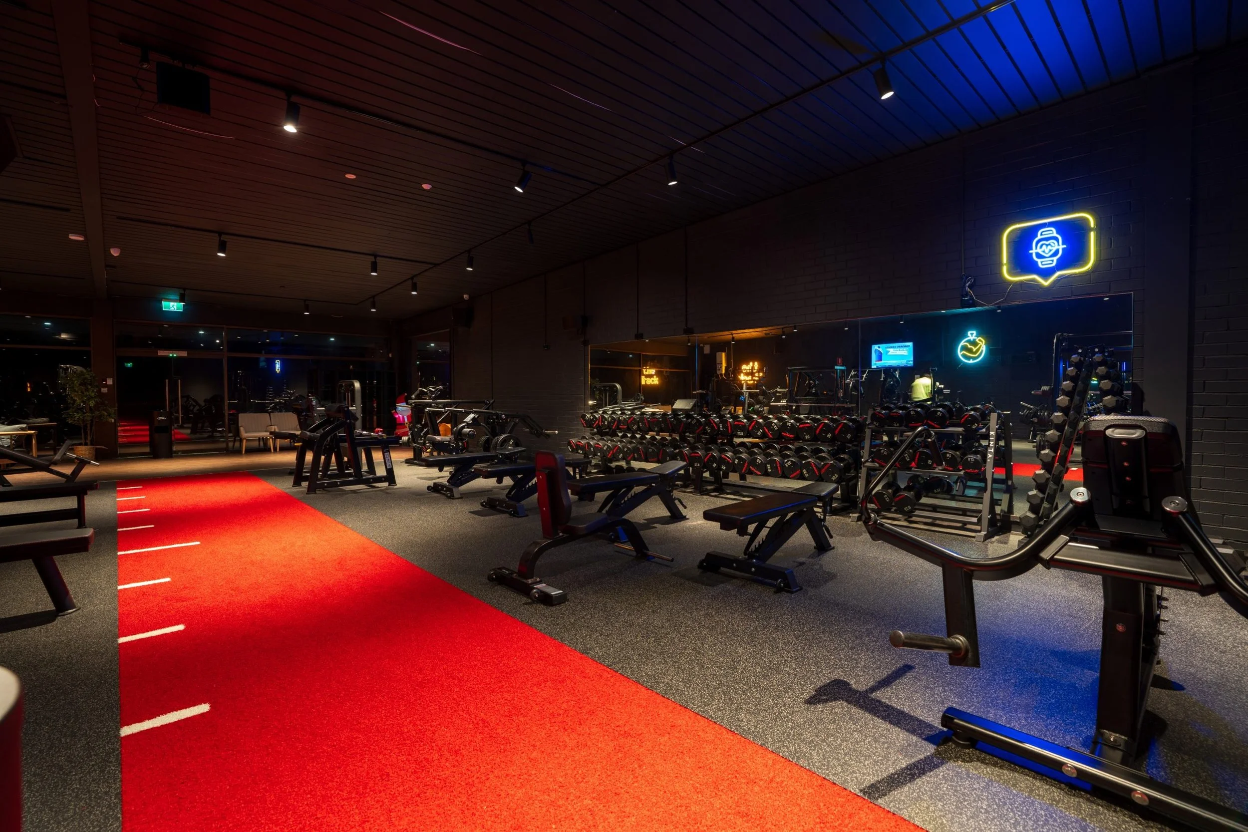 A modern gym with black brick walls, a red running track, and various equipment including dumbbells, benches, and cardio machines. Neon signs glow on the walls, and ceiling lights illuminate the space. Large windows are visible along one wall.