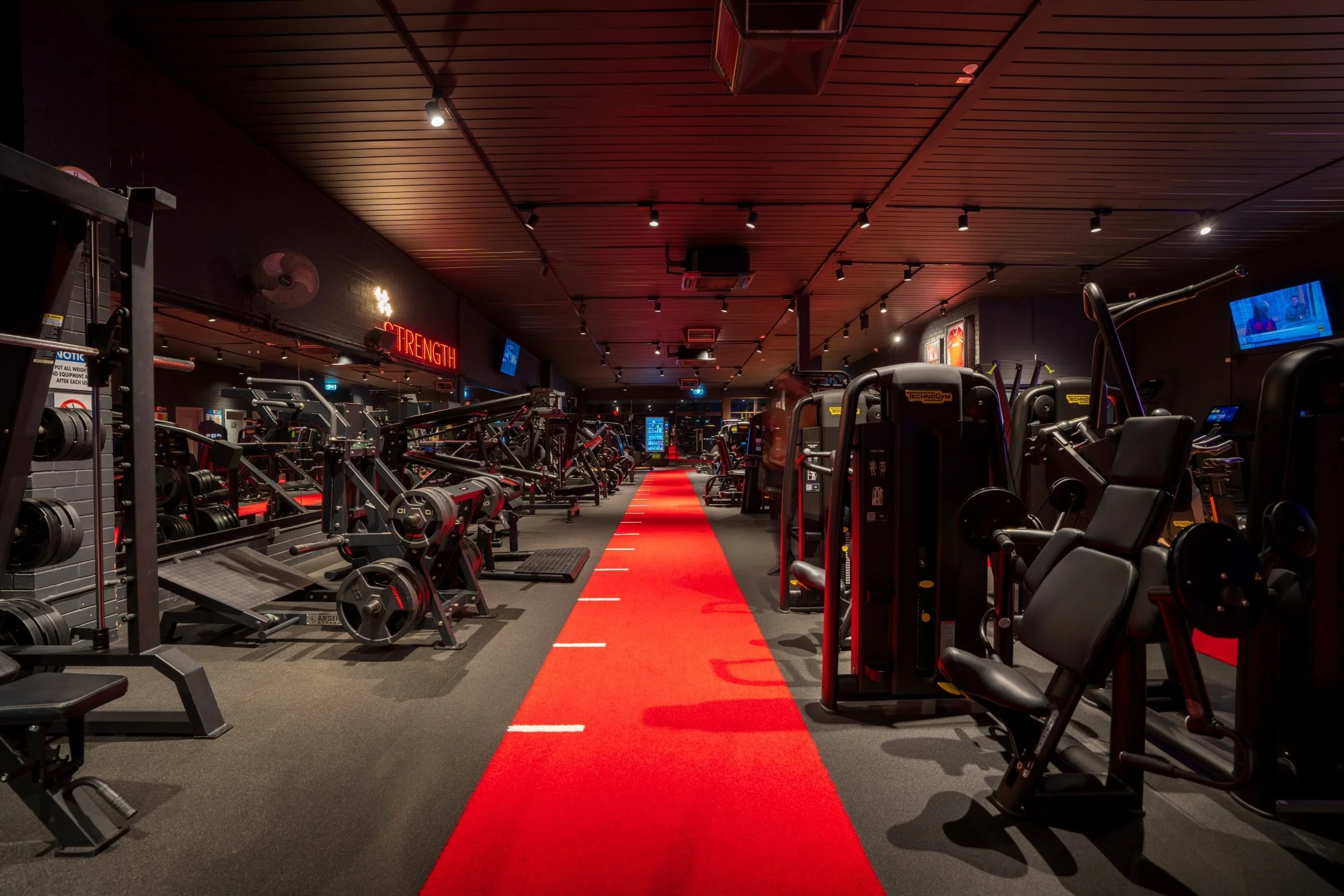 Interior of a modern gym with black walls and a ceiling with wooden panels. The gym has various workout equipment including weight machines and free weights. A red carpet runs down the center of the gym, and the word 'STRENGTH' is illuminated in red 