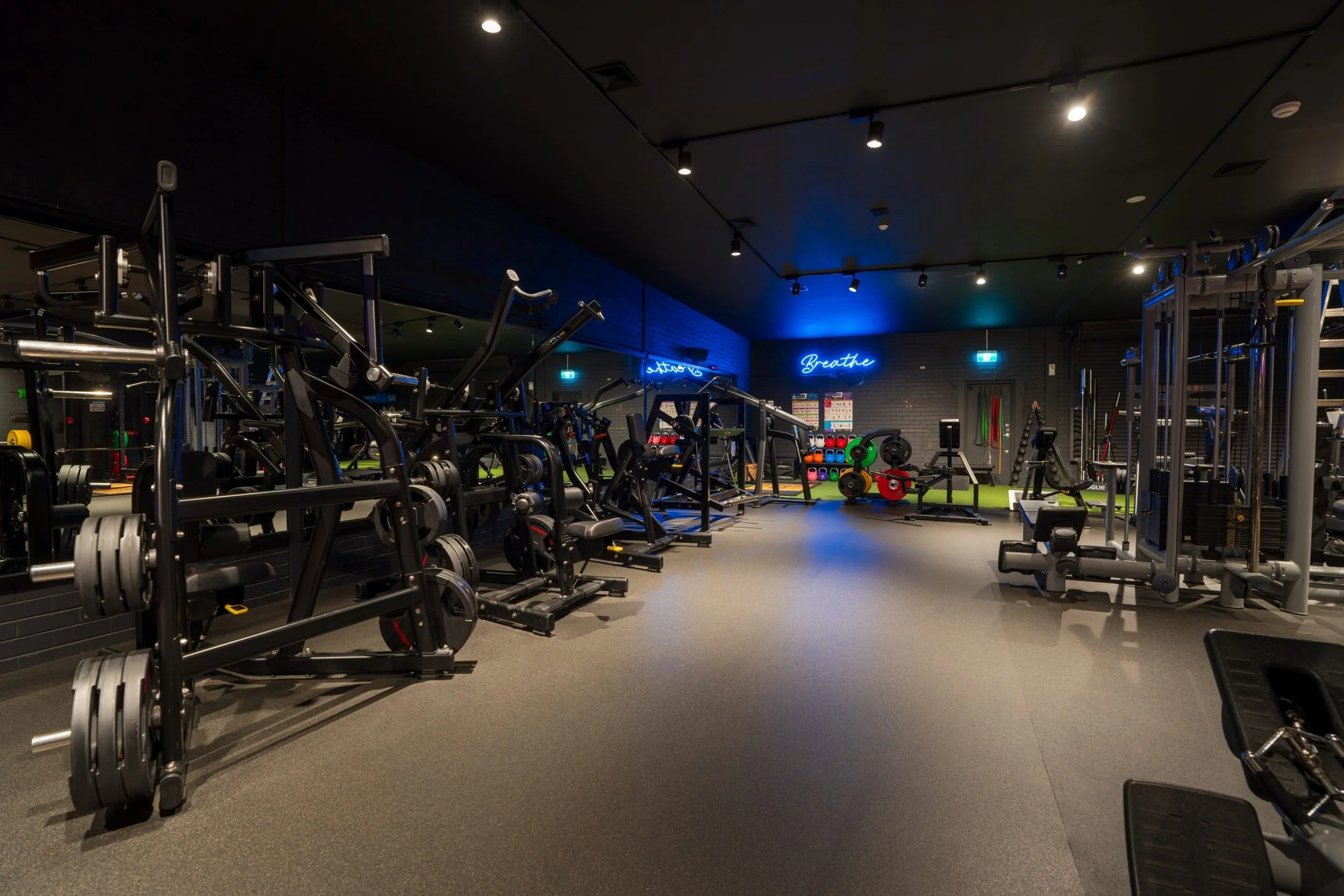An interior of a modern gym with various weightlifting machines, free weights, and exercise equipment. The space is dimly lit with overhead lighting and a blue neon sign that says 'Breathe' on a brick wall.