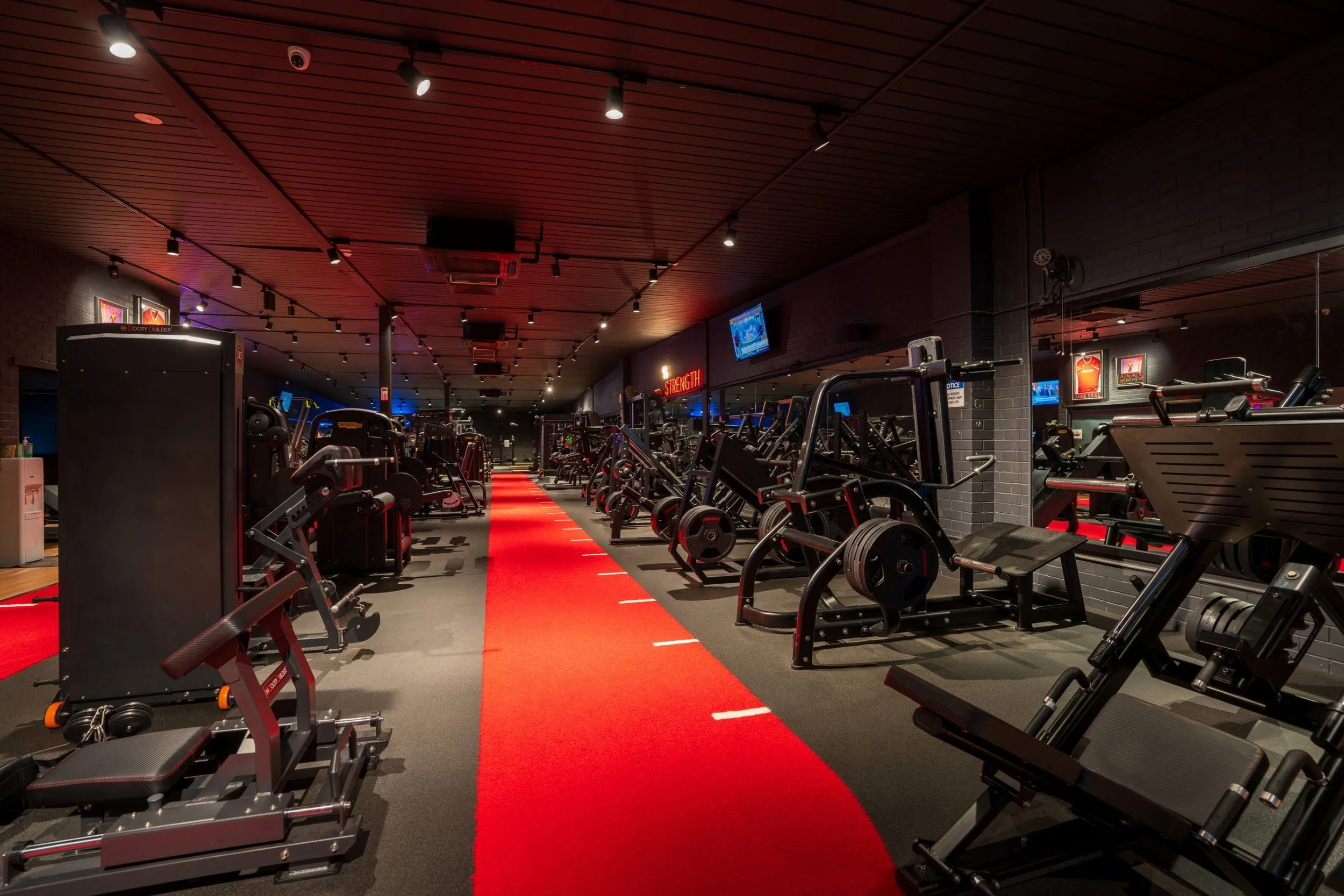 Gym interior with various workout equipment including weights, cardio machines, and a red carpet running through the center under ceiling lights.