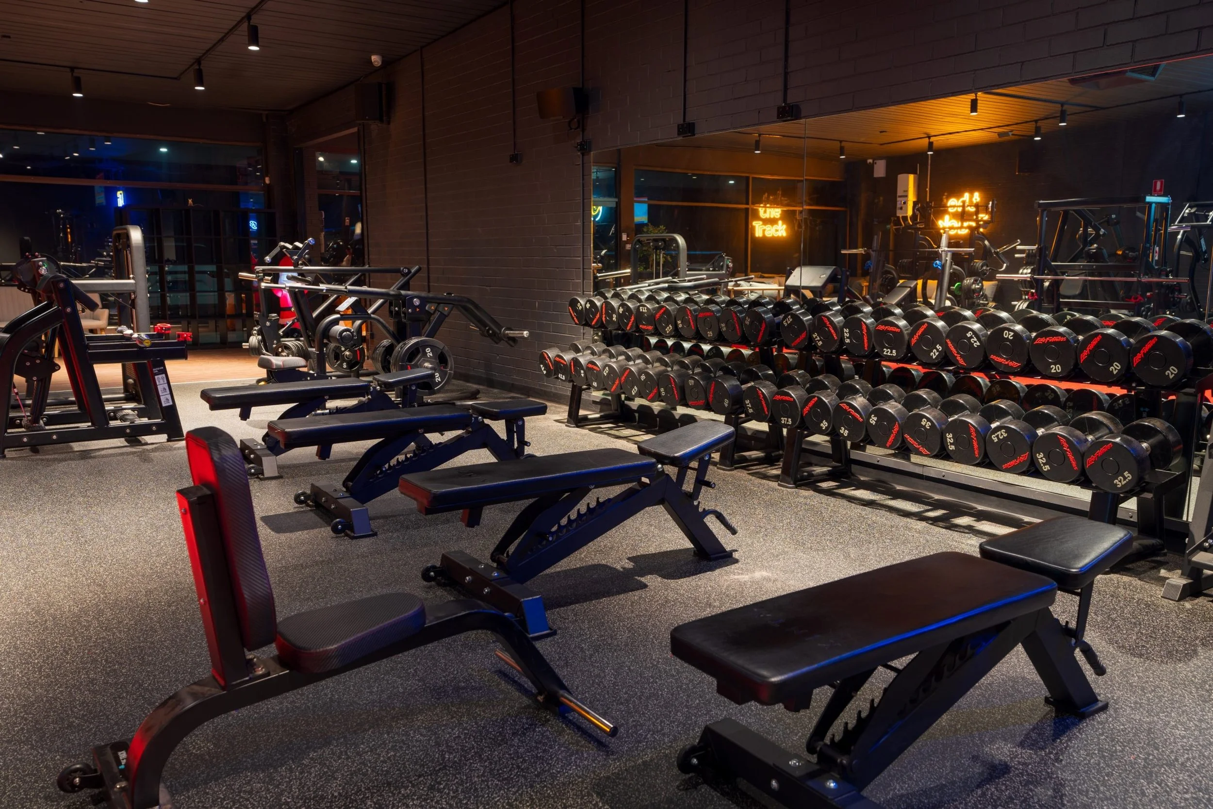 A gym with various workout equipment, including dumbbells on a rack and exercise benches, illuminated by focused ceiling lights and surrounded by large windows reflecting neon signs outside.