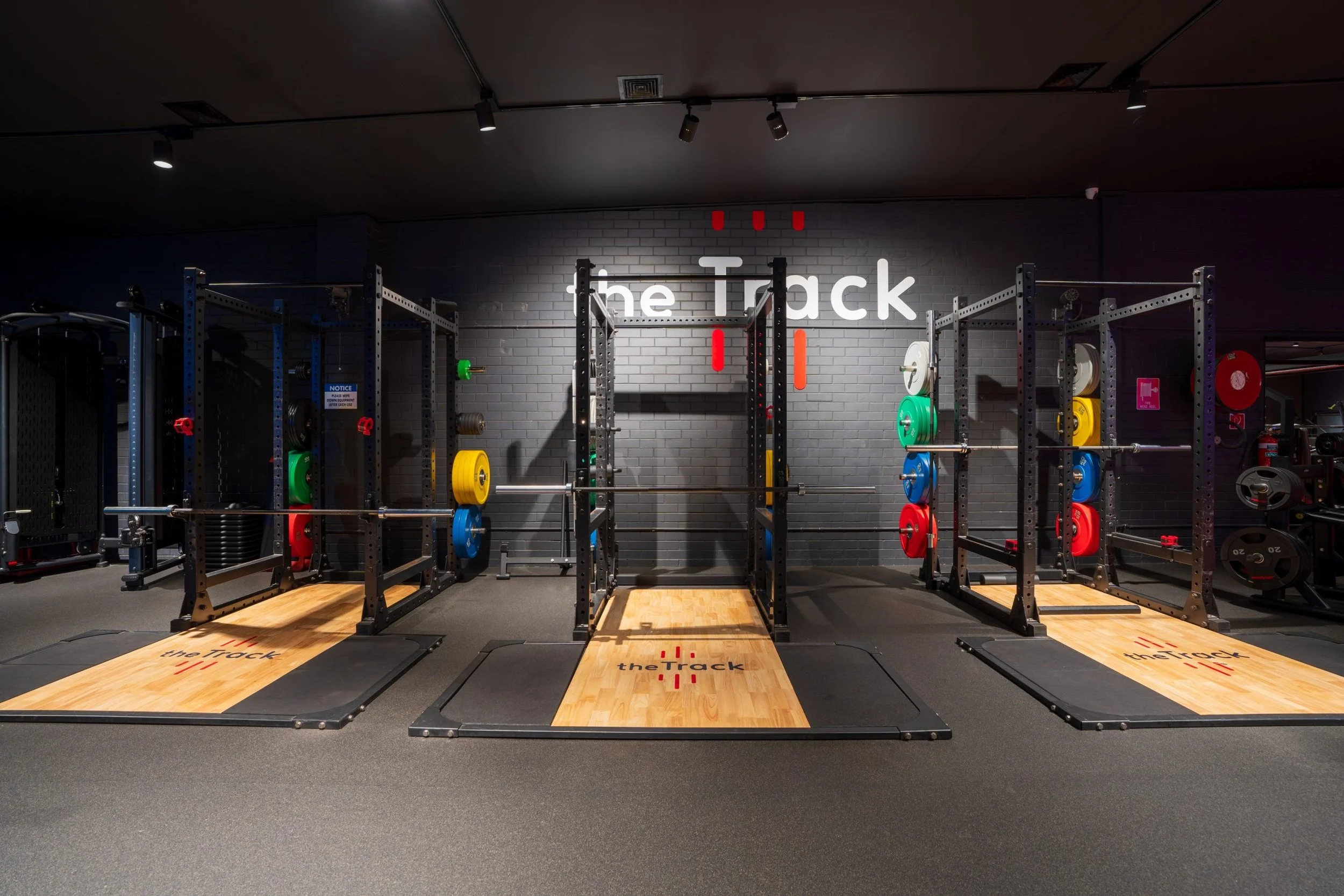 A modern gym with black metal squat racks and weight plates in various colors, including green, yellow, red, and blue, arranged on the racks. The floor features wooden platforms with the words 'the Track' written on them. The background has a gray br