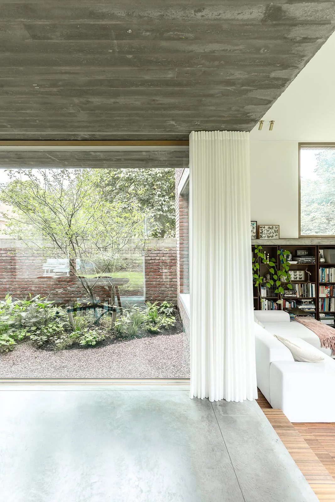 Binnenruimte met grote glaswand die naar een tuin uitkijkt, boekenkast, witte bank, en witte scheidingsgordijn.