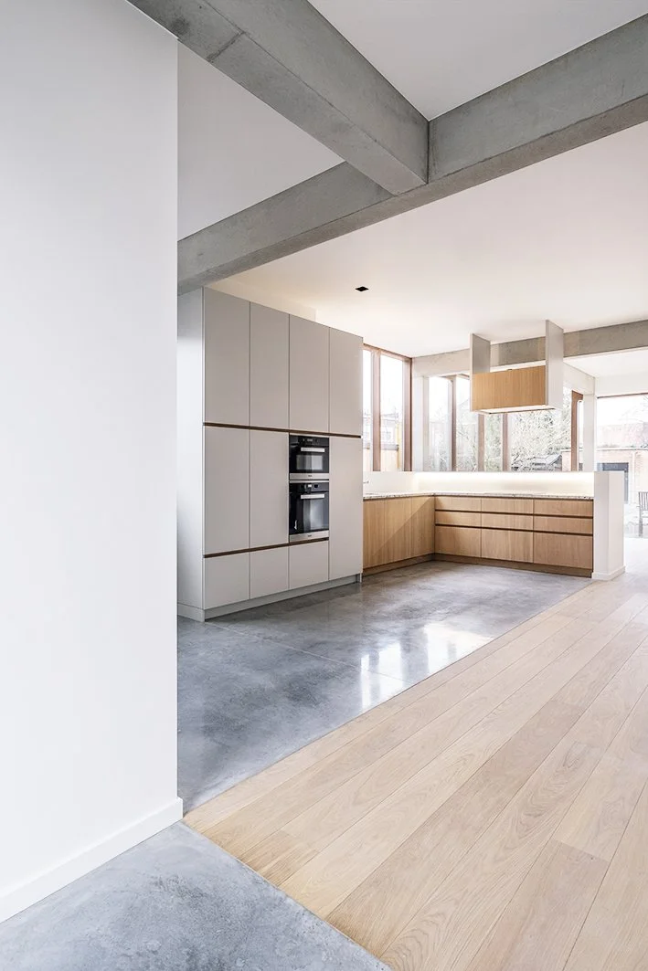 Moderne keuken met houten en glazen kasten, ingebouwde oven, grote ramen en een mix van betonnen en houten vloeren.