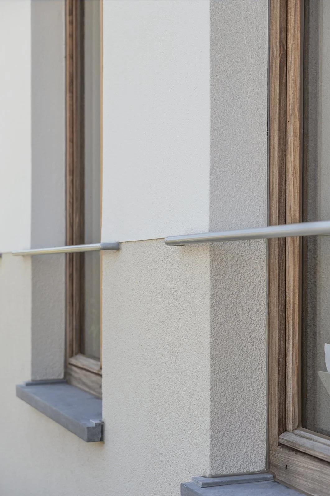 Buitenkant van een huis met een raam en een houten deur, een metalen handrail en een lichtgekleurde muur.