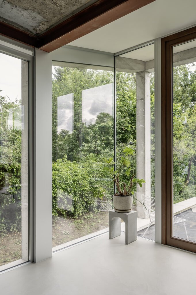 Interieur van een modern huis met grote glazen ramen, groene tuin buiten, een plant op een klein wit tafel, en een witte stoel buiten te zien.