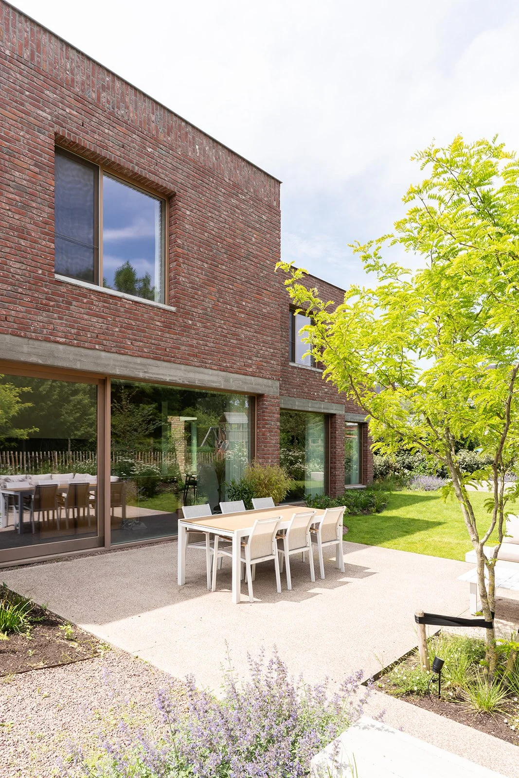 Uitzicht op een moderne woning met grote glazen deuren en ramen, een tuin met een tuinmeubelset, en een boom met groene bladeren.
