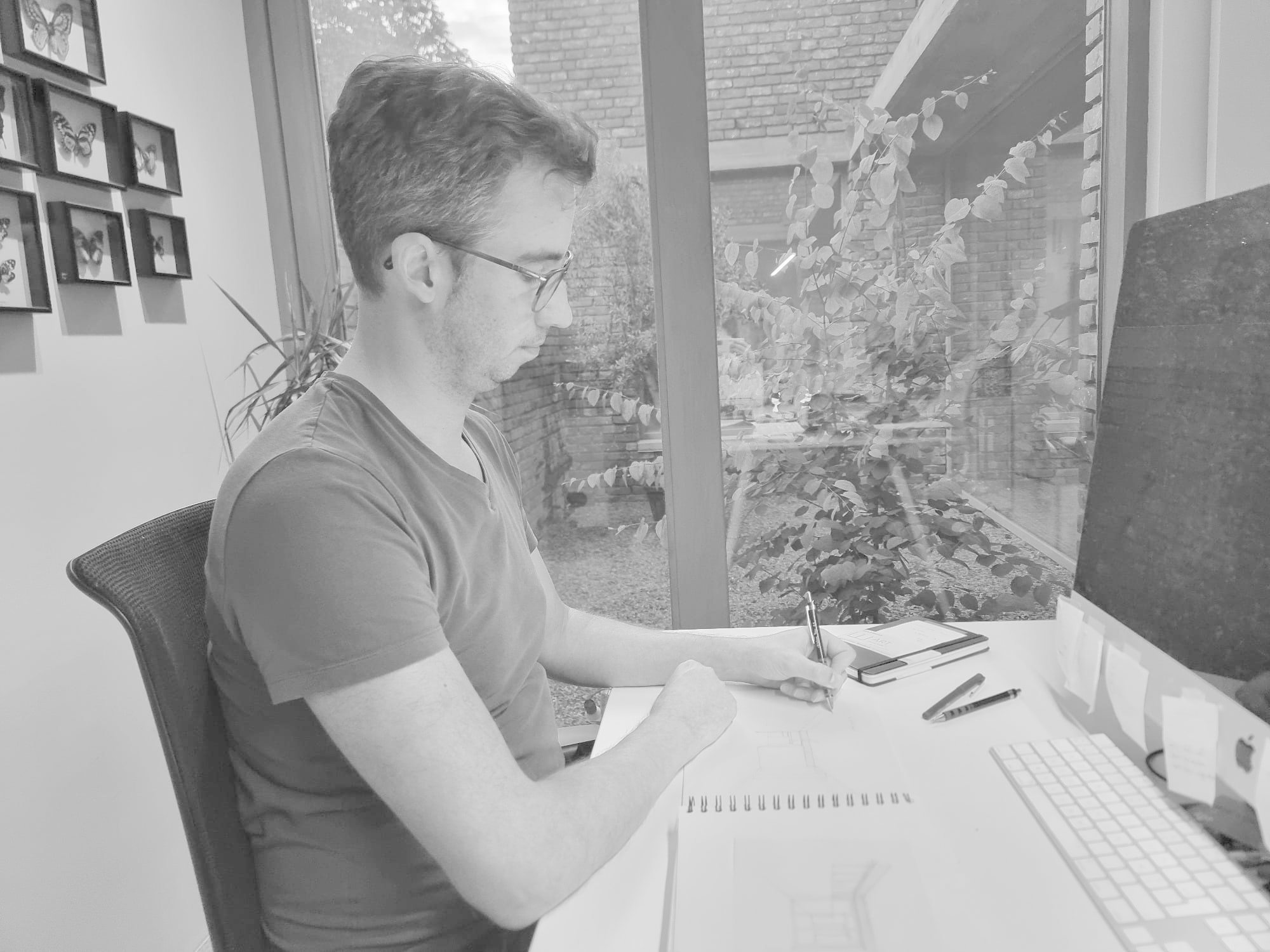 Man zit aan een bureau met een laptop, papier en pennen, terwijl hij naar buiten kijkt door een groot raam, in een kantoor met muurdecoraties.