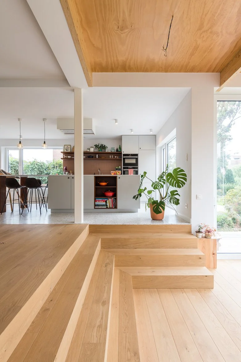 Een interieur van een moderne woning met een houten trap, een open keuken met witte kasten en een kookeiland, grote ramen met uitzicht op de tuin, en groene planten.