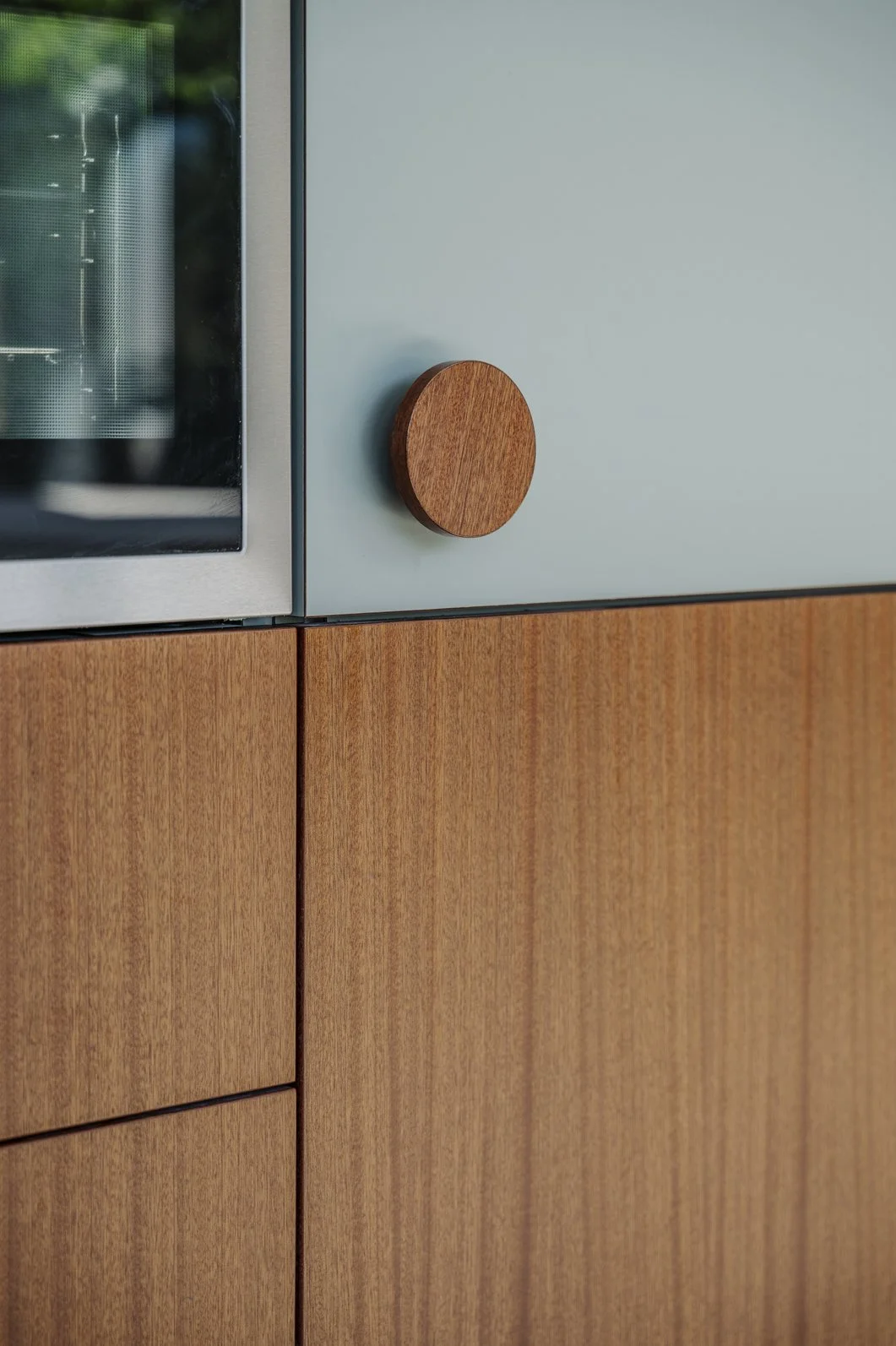 Close-up van een keukenkast met houten panelen en een ronde houten knop, naast een oven met een glazen deur.