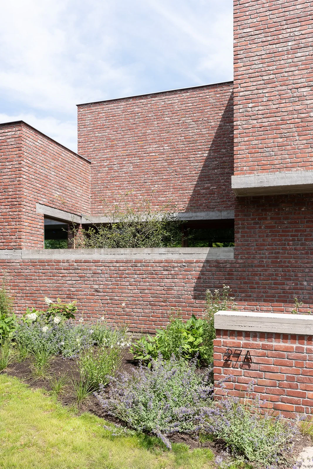 Een modern gebouw met rode bakstenen en grote open ramen, omringd door een tuin met bloemen en groen.