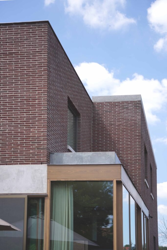Modern gebouw met rood bakstenen muren, grote ramen met groene gordijnen, en een heldere blauwe lucht met wat wolken.