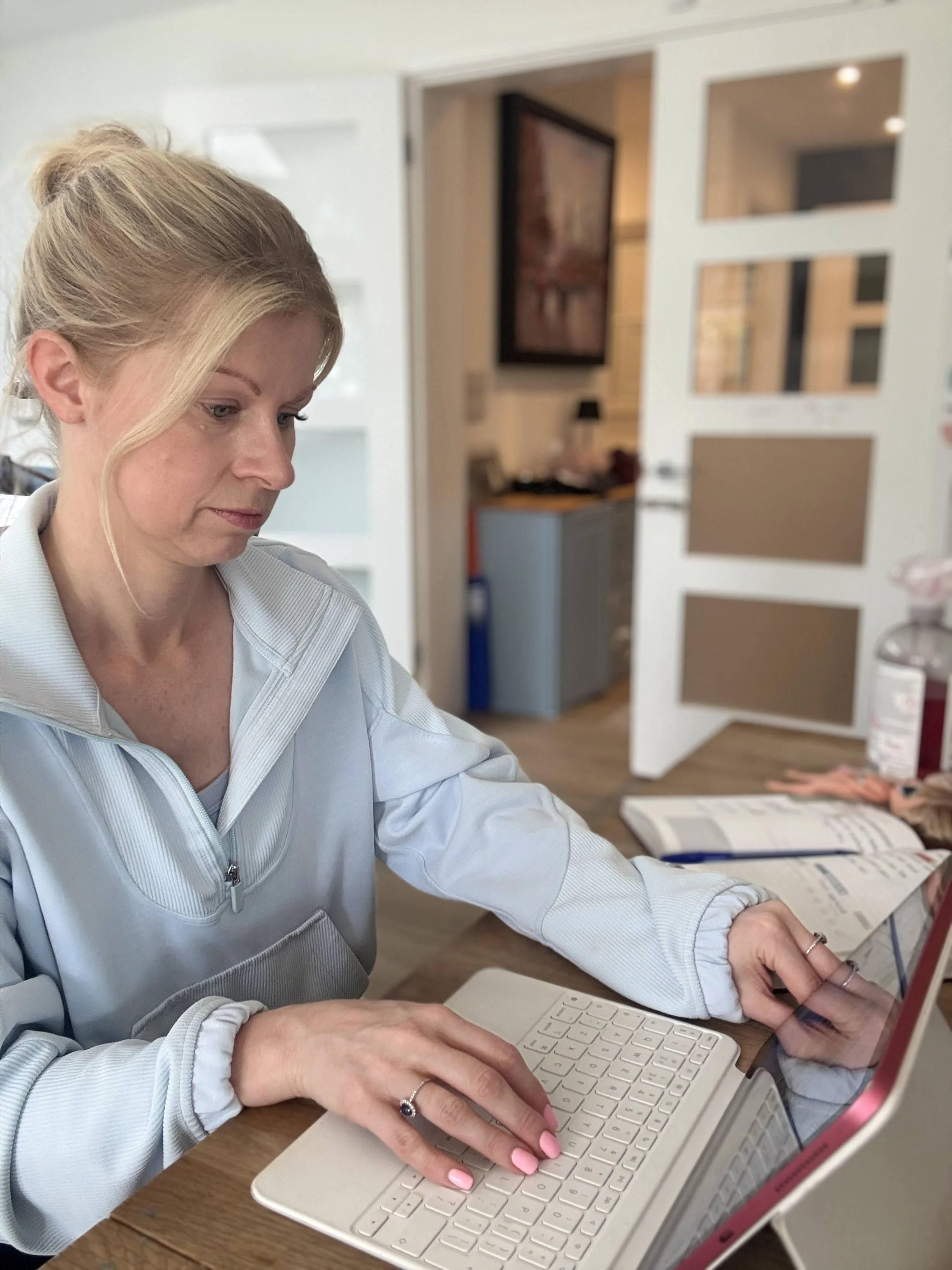 A woman with blonde hair in a ponytail is typing on a white keyboard connected to a laptop with a red frame. She is seated at a wooden table in a room with an open door and house decor visible in the background.