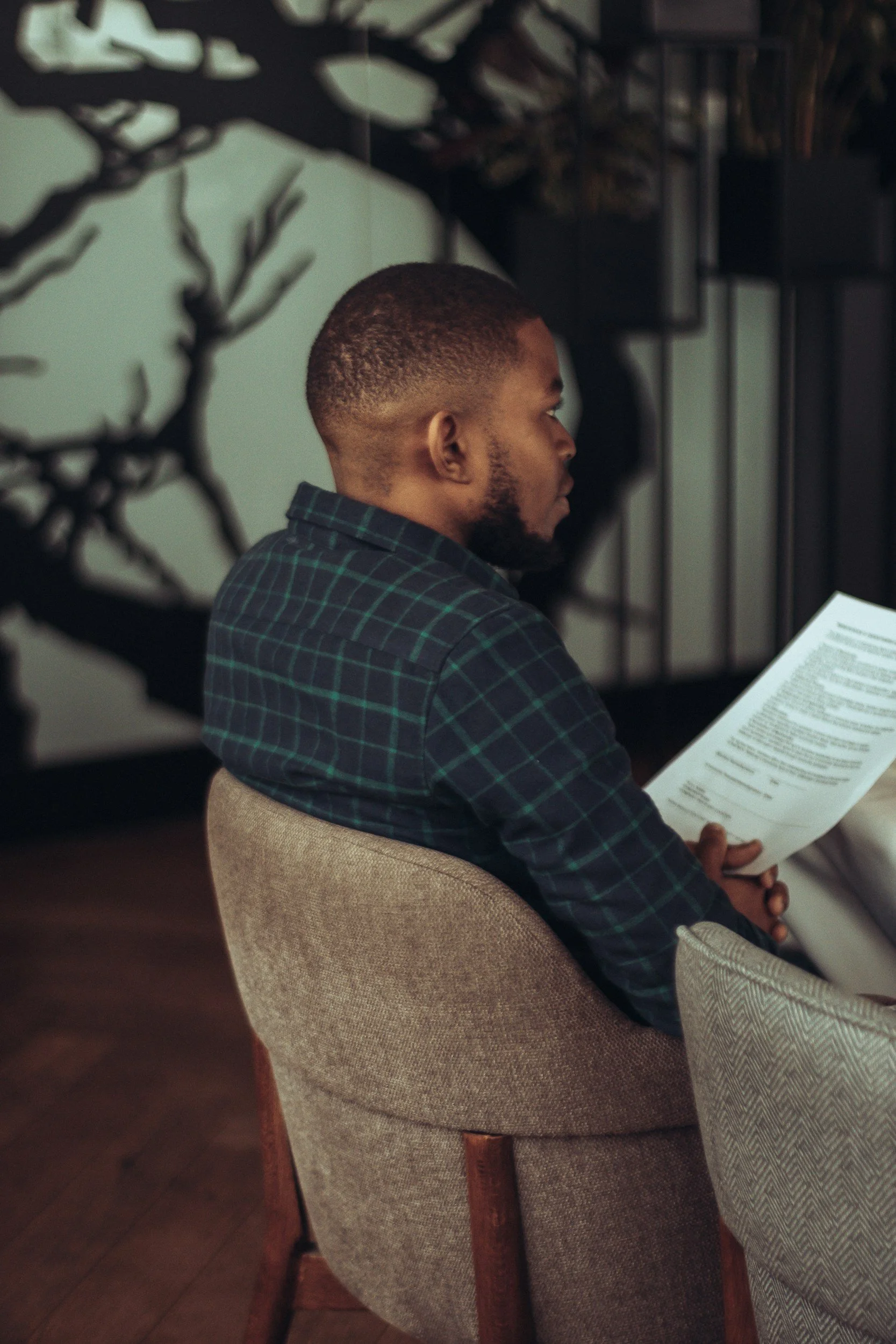 A young man with short hair and a beard, wearing a dark plaid shirt, sitting on a beige chair and reading a document. The background includes a dark wall with tree branch and leaves silhouette art decor.