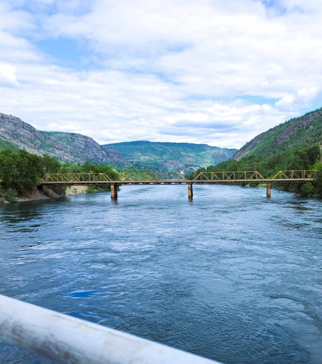 View of a river flowing between green hills with a metal bridge across the water and cloudy sky overhead.