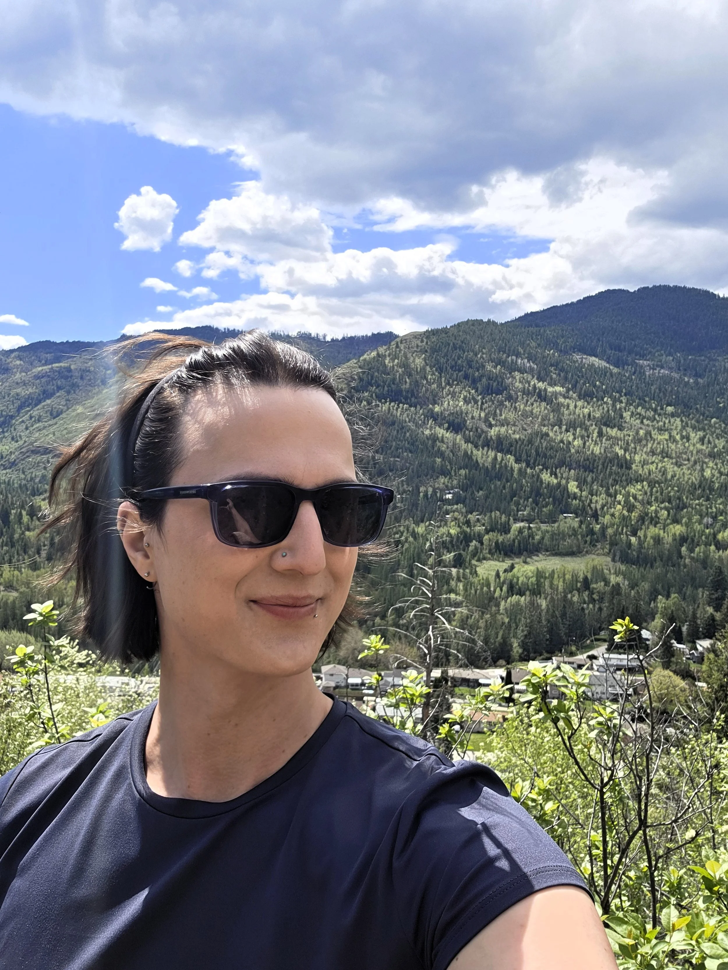 A person with short dark hair, wearing sunglasses and a black shirt, smiling in front of a mountainous landscape with scattered clouds in the sky.