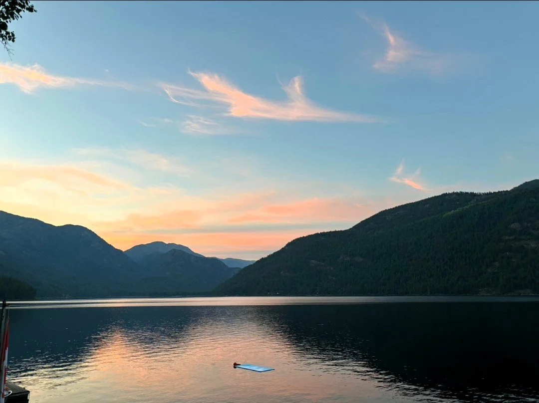 A calm lake surrounded by green mountains under a colorful evening sky with pink and blue clouds.