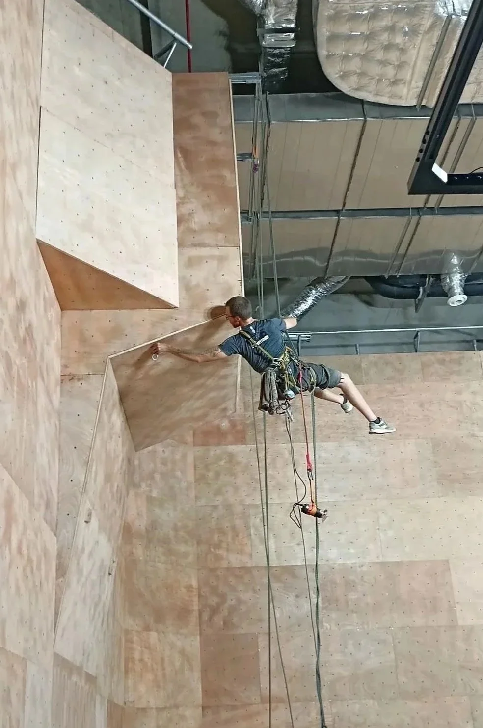 A person in climbing gear cleaning the ceiling of an indoor rock climbing gym.
