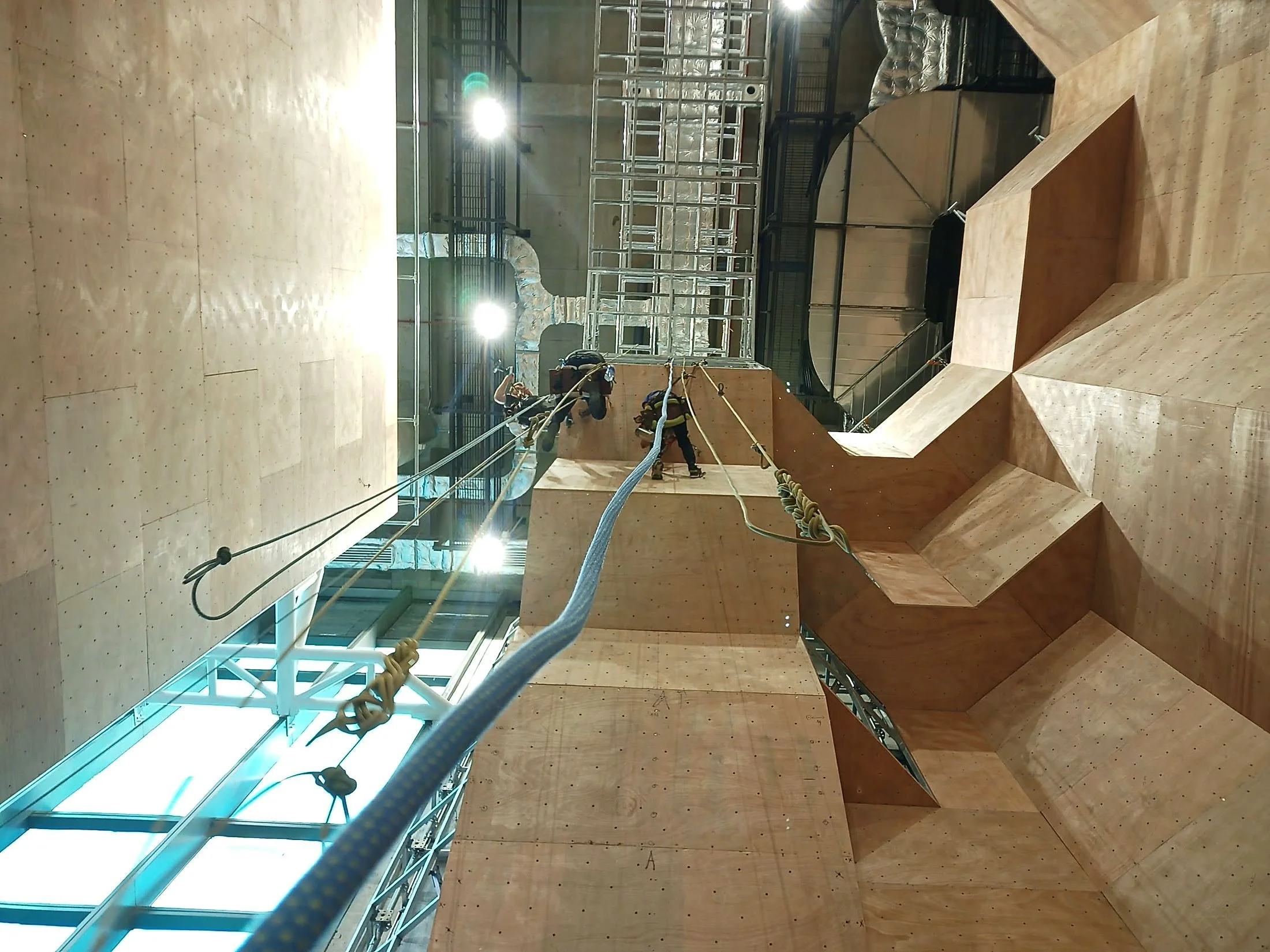 Inside a modern building under construction or renovation, showing a high ceiling, wooden stairs, and some workers installing or repairing infrastructure. There are ropes, safety harnesses, and construction equipment visible.