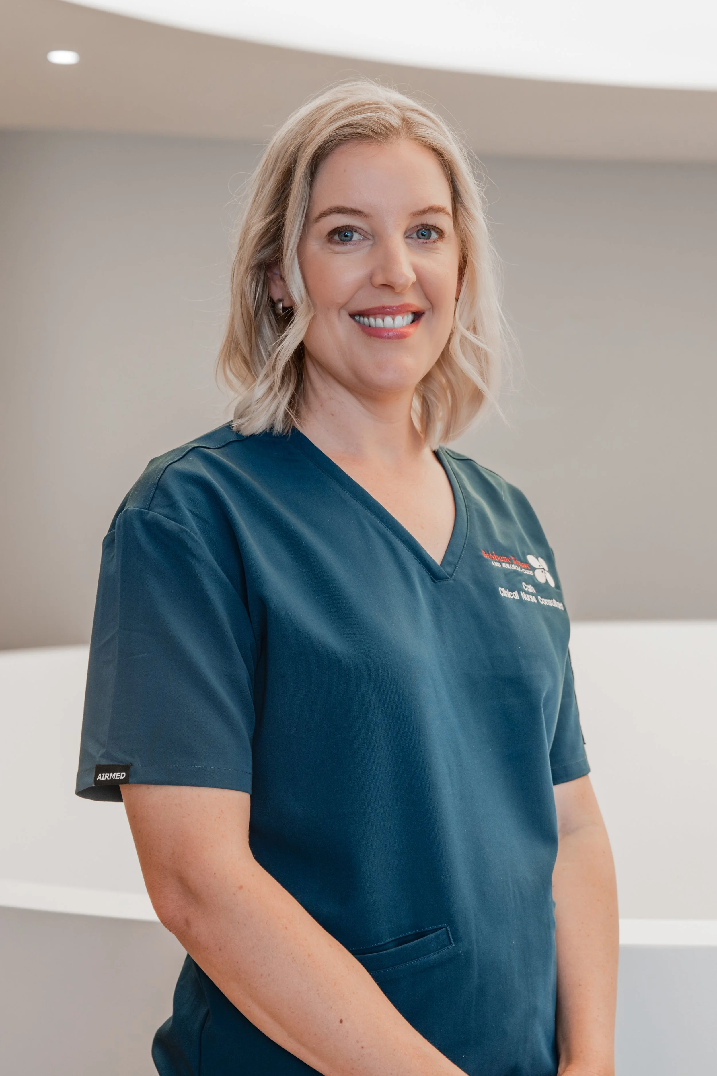 A woman with blonde hair wearing blue scrubs with a hospital logo, standing in a clinical setting, smiling at the camera.