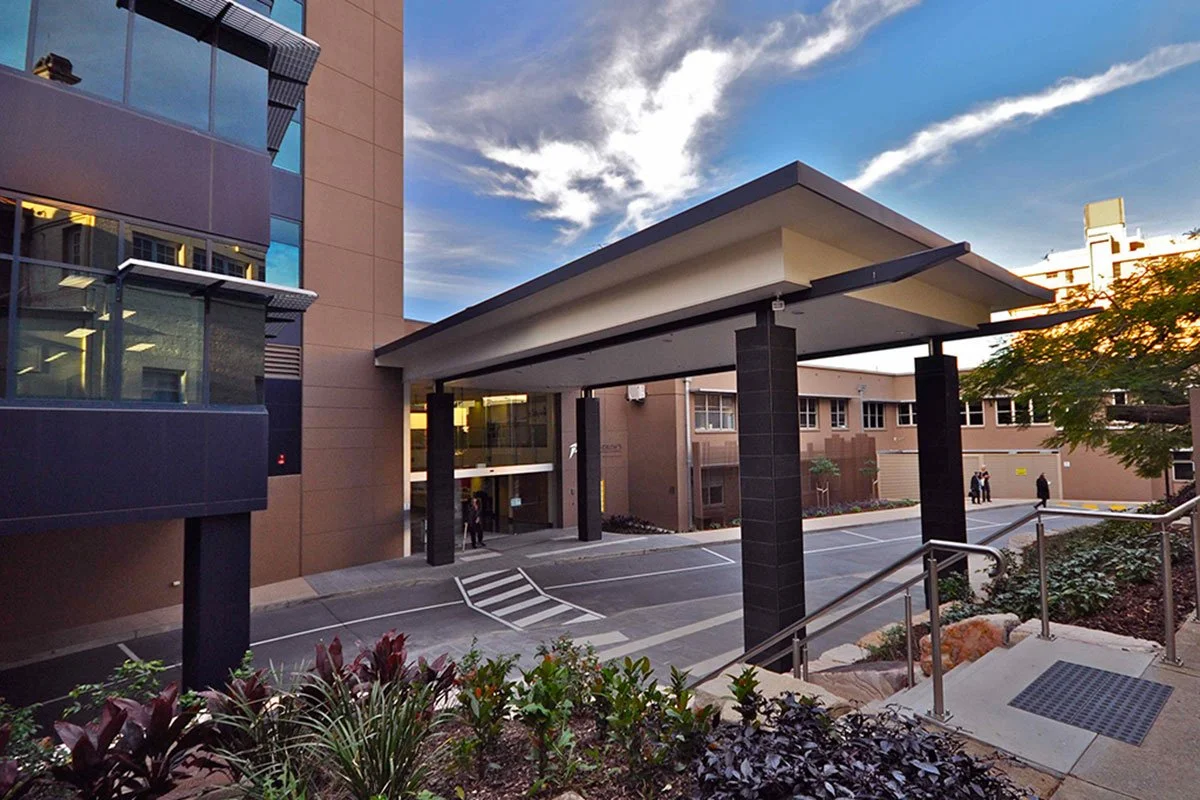 View of a modern office building entrance with a covered driveway, black columns, a ramp, and landscaped plants in the foreground, under a partly cloudy sky.