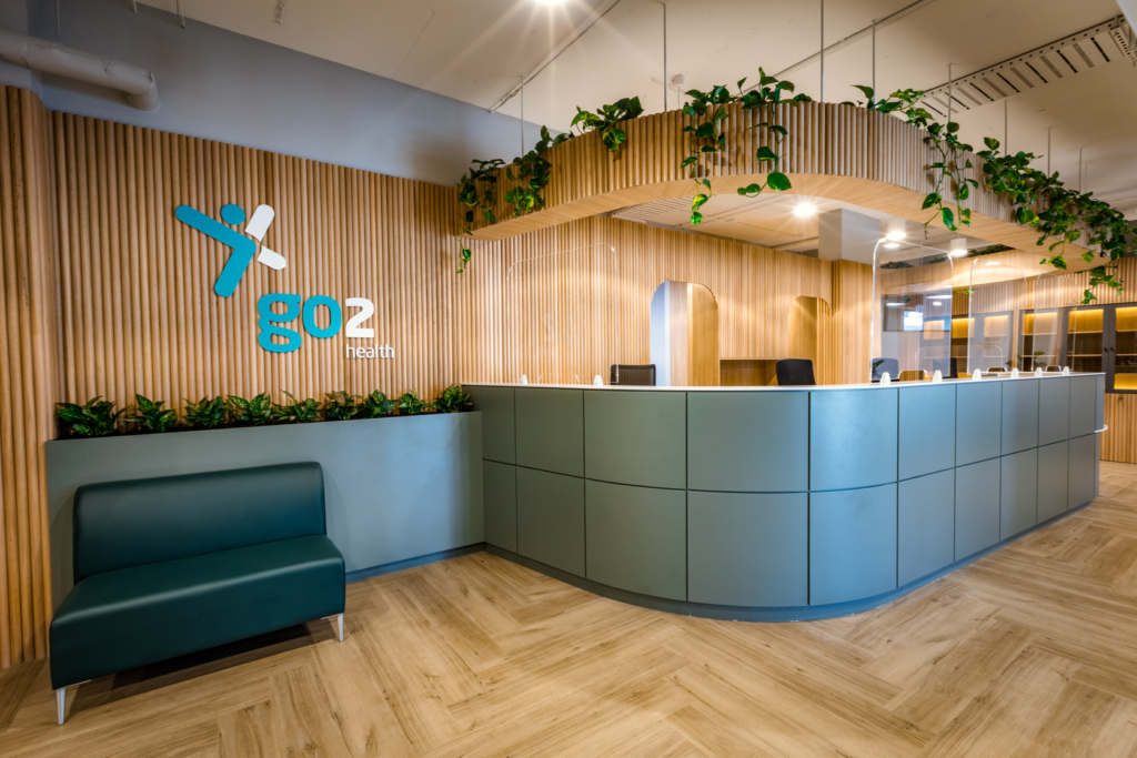Modern reception area with a curved desk, green panels, a teal bench, wood accents, and green plants, with the logo 'go2 health' on a wooden wall.