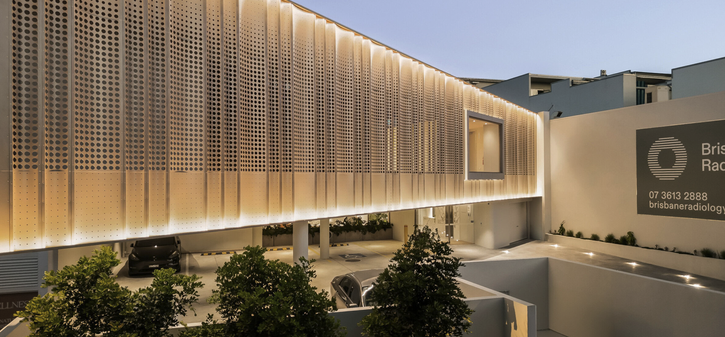 Modern building facade with perforated metal panels, illuminated lighting, and a parking area below, with nearby trees and a large sign displaying contact information for Brisbane Radiology.