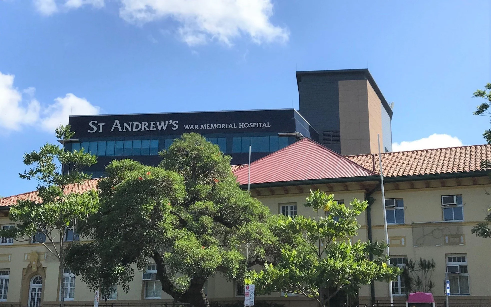 The image shows a building with a sign that reads 'St Andrew's War Memorial Hospital'. The building has a modern glass section on top and a traditional section with a tiled roof. There are trees in the foreground and a clear blue sky with some clouds overhead.