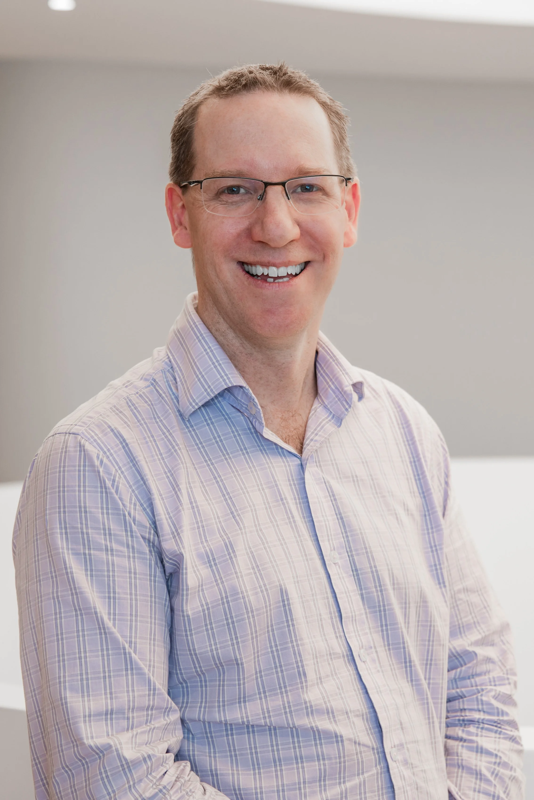 A smiling man with glasses wearing a light-colored checkered shirt.