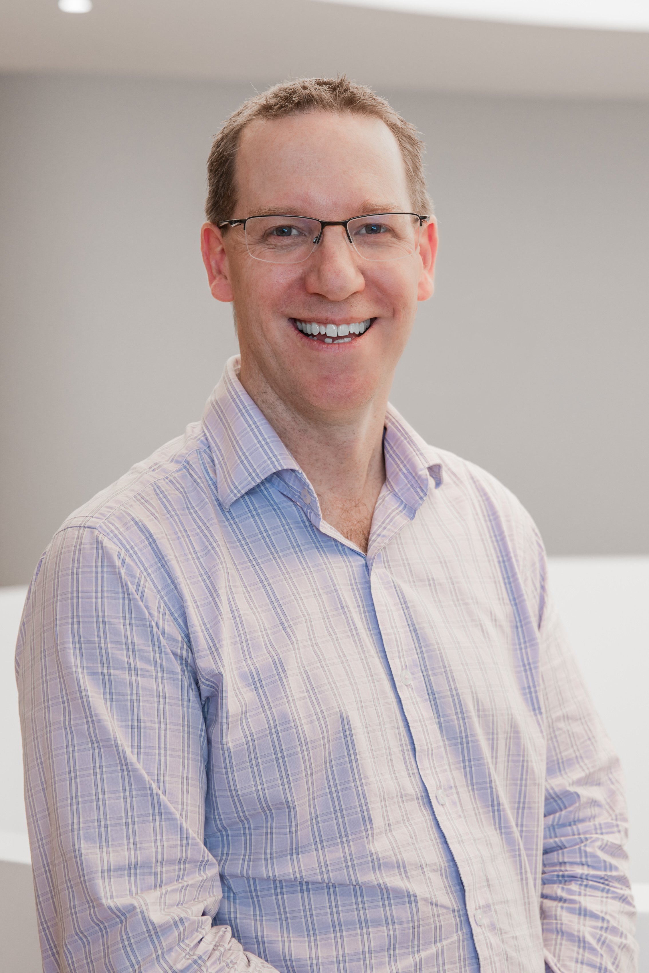 A smiling man with glasses wearing a light purple plaid shirt, standing indoors with a plain light-colored wall behind him.