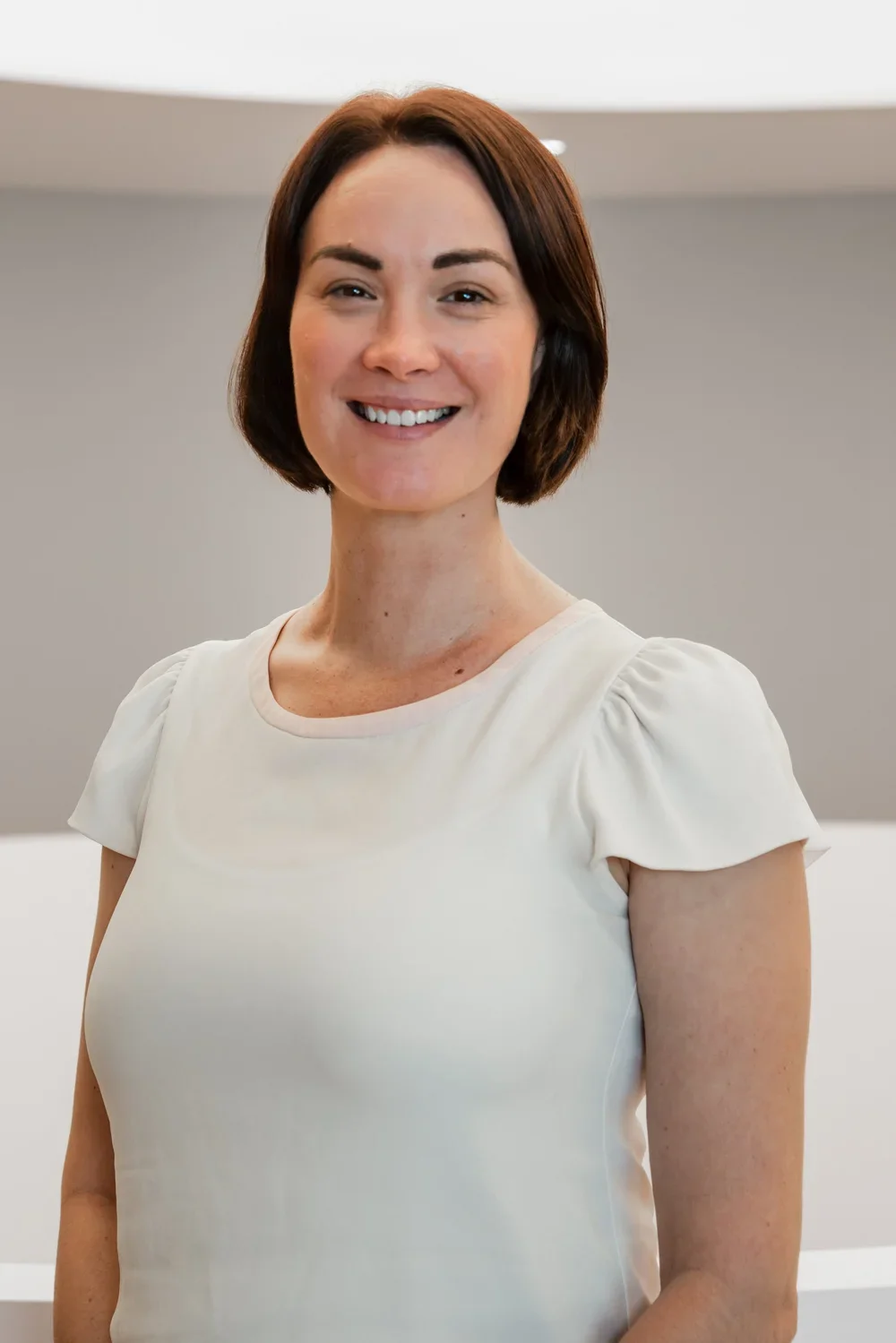 A smiling woman with short brown hair wearing a white top, standing indoors.