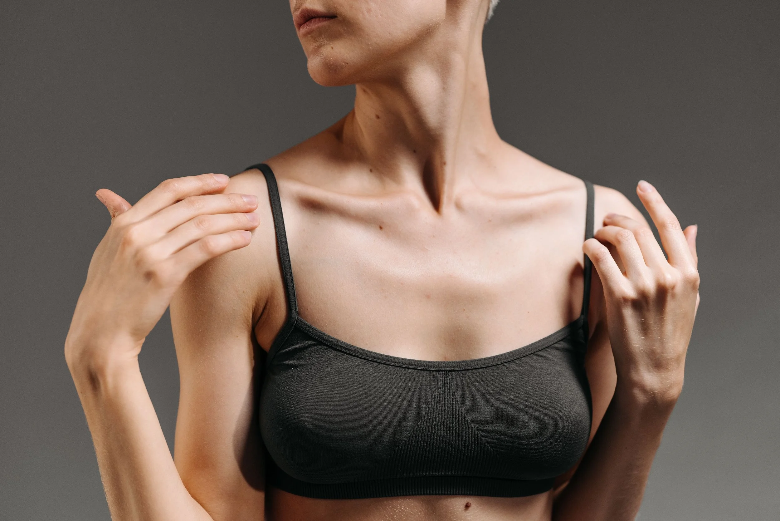 Close-up of a woman wearing a black tank top, with her hands resting on her shoulders, against a dark gray background.