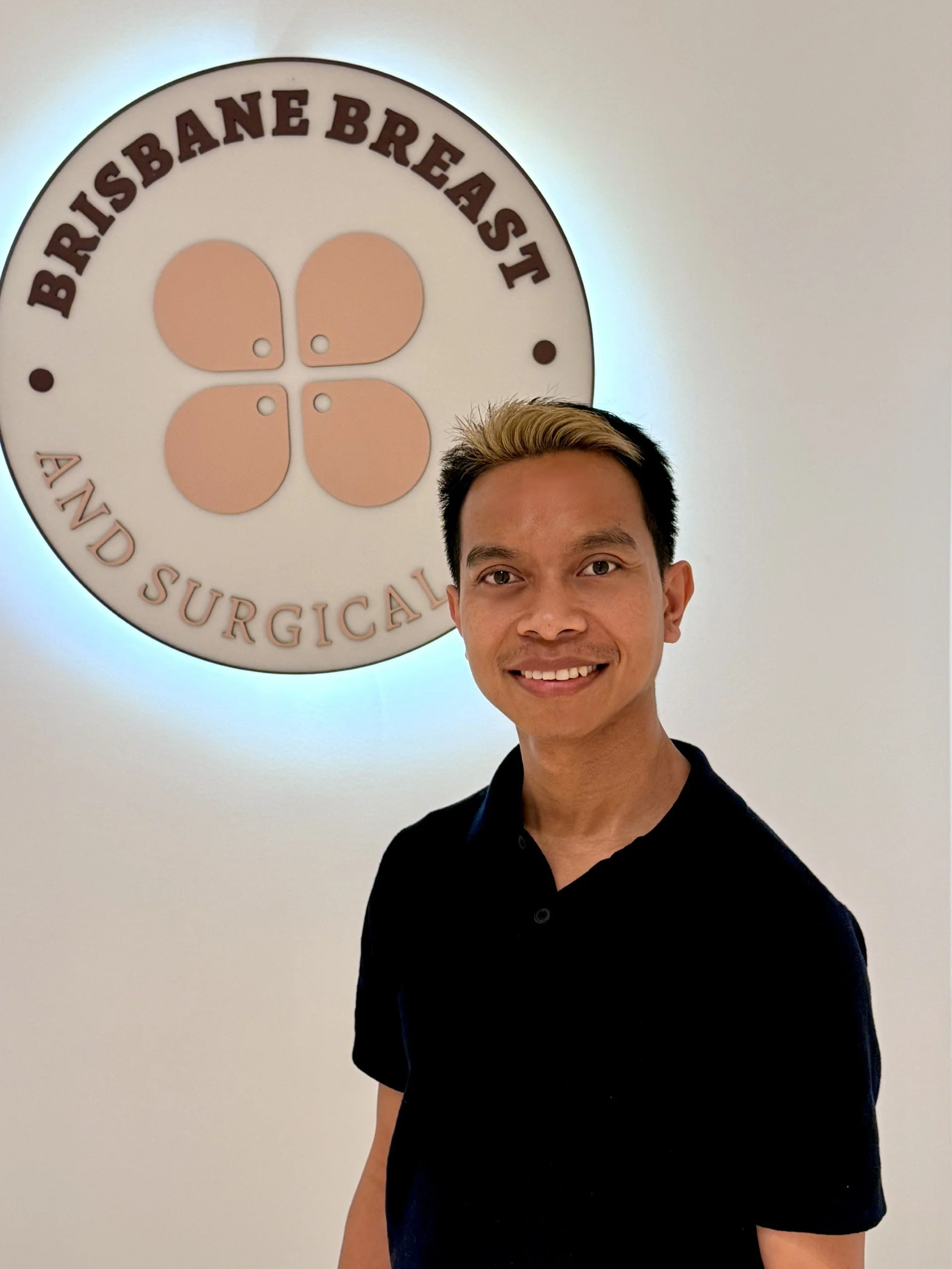 Young man smiling in front of a circular sign with the text 'Bristbane Breast' and an illustration of four rounded shapes with small holes, on a white wall.