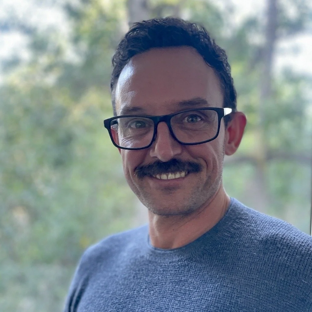 Smiling man with glasses and a mustache wearing a blue shirt, standing outdoors with trees in the background.