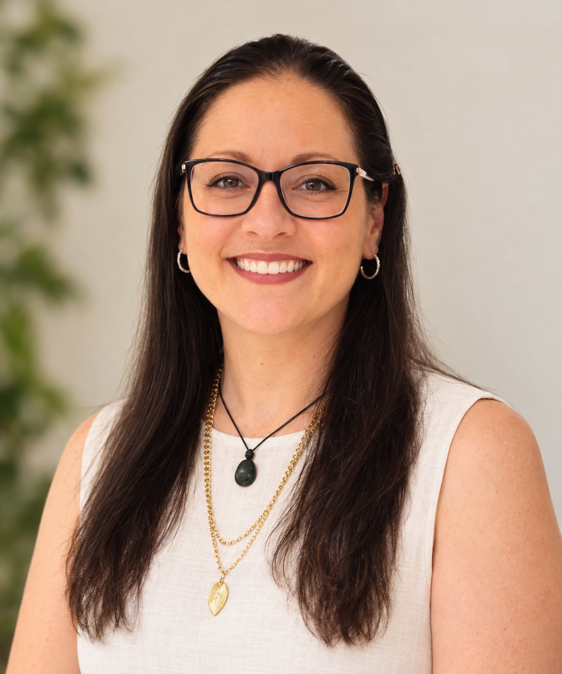 A woman with long dark hair, wearing glasses and earrings, smiling, dressed in a sleeveless white top, with layered necklaces, including a black pendant and gold leaf pendant, with a blurred green plant in the background.